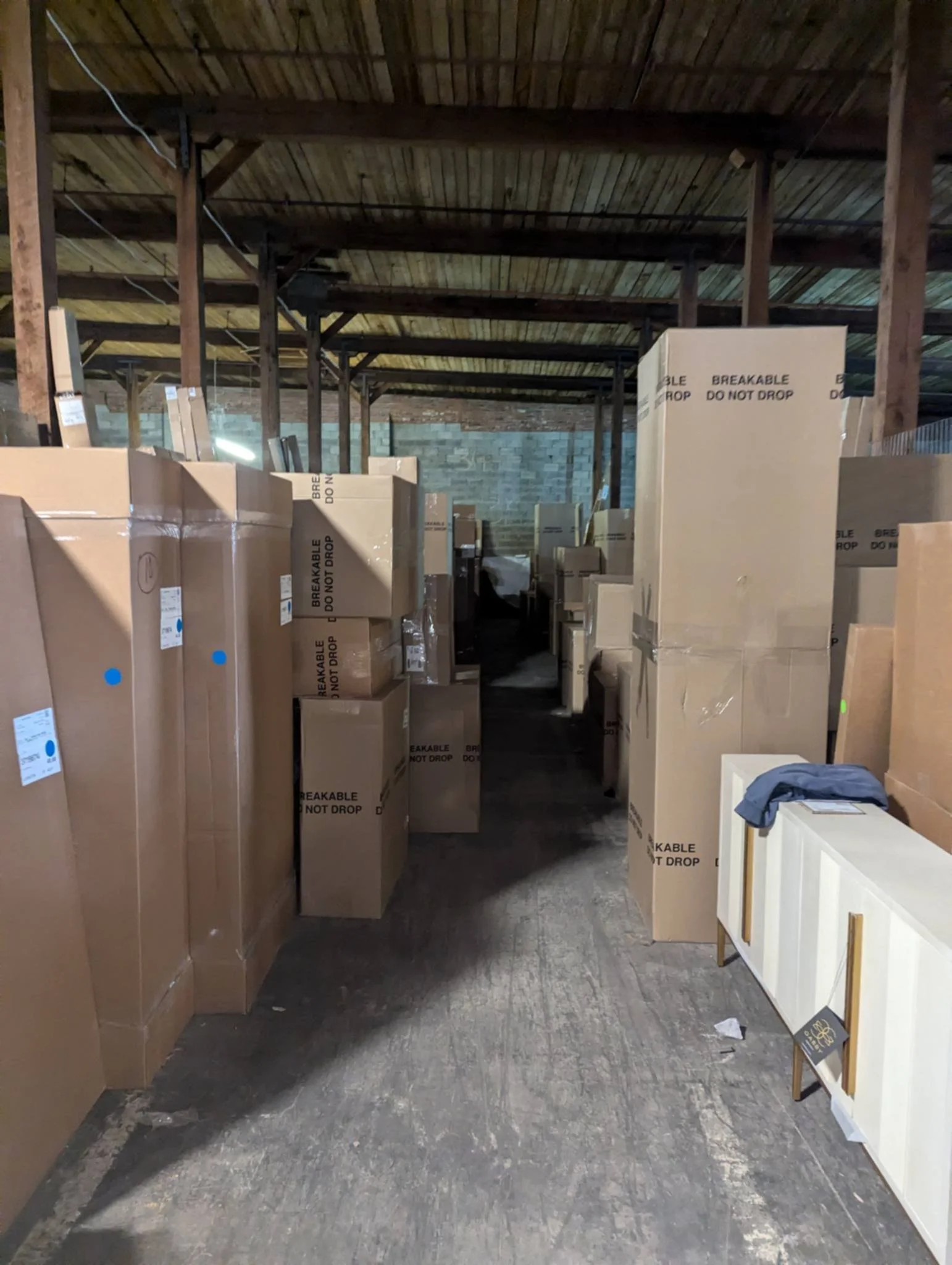 Warehouse aisle with tall cardboard boxes and furniture, including white cabinets, stored on a dark wooden floor under a wooden ceiling.