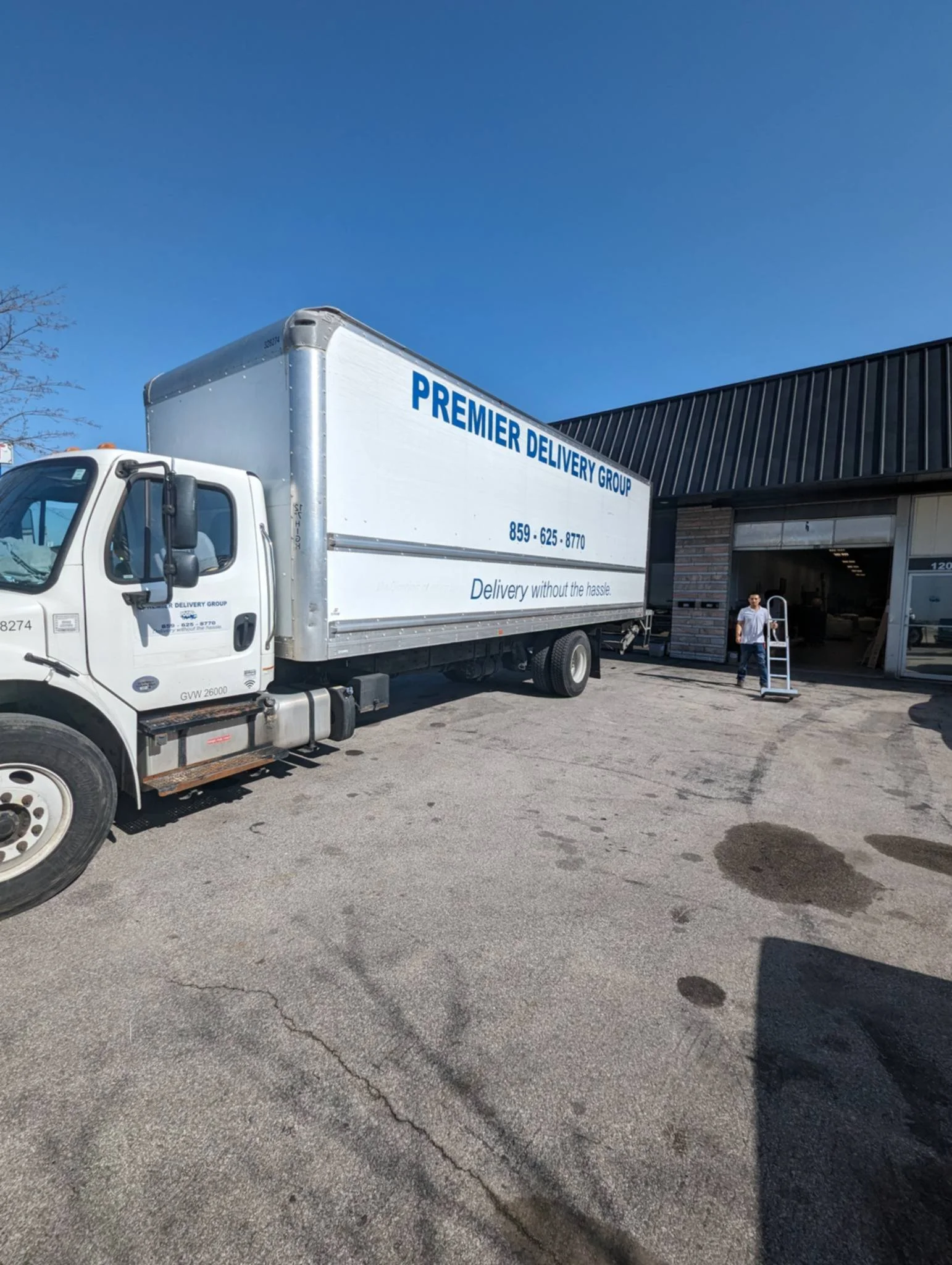 A white delivery truck branded 'Premier Delivery Group' parked outside a building, with a person holding a dolly near the entrance on a sunny day.