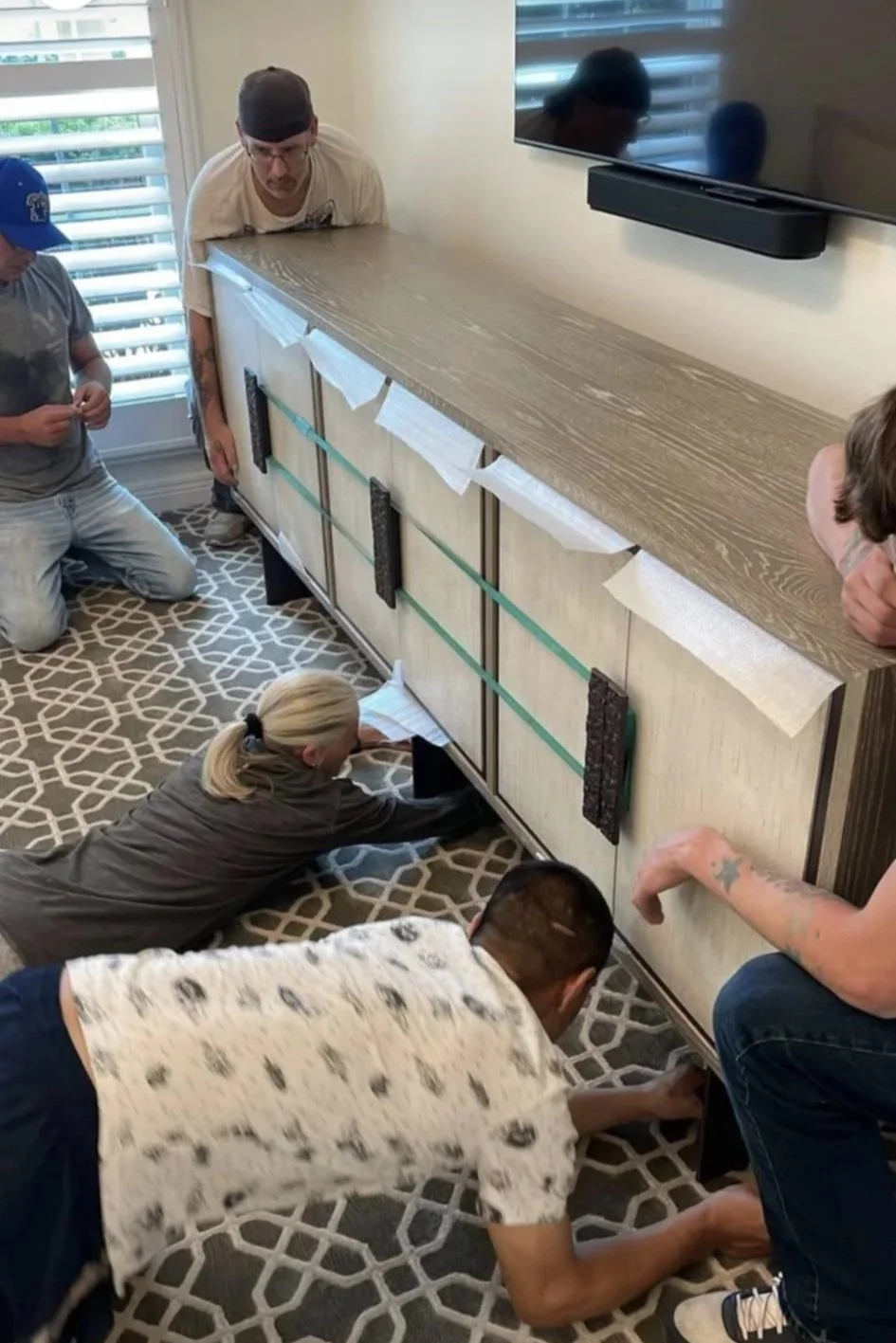 People working together on a furniture project in a room with a TV on the wall and a window with shutters.