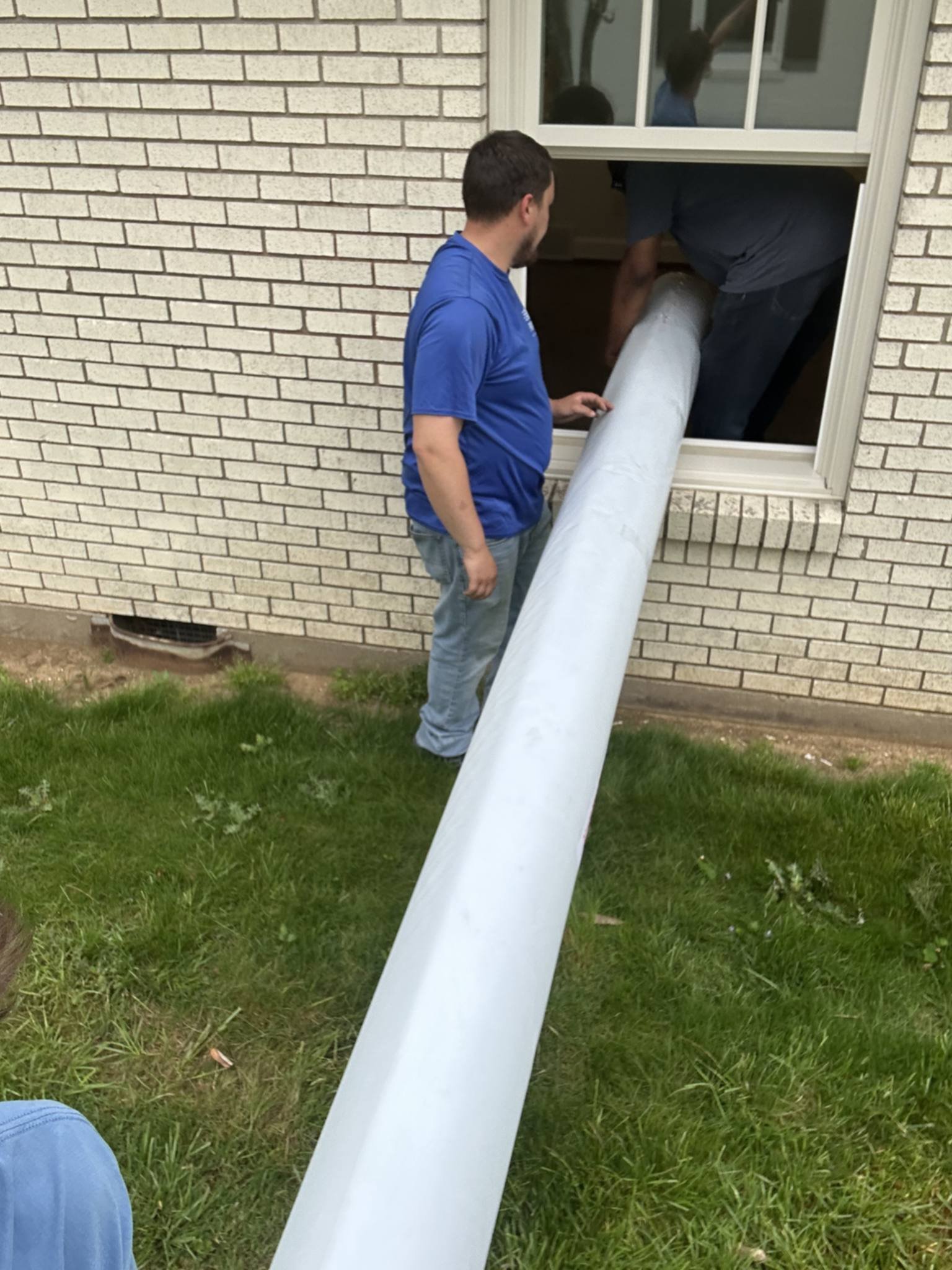 Two men are installing a large white funnel into a house window. One man is standing outside near the grass and the other is inside the house, both handling the funnel as it goes through the window.