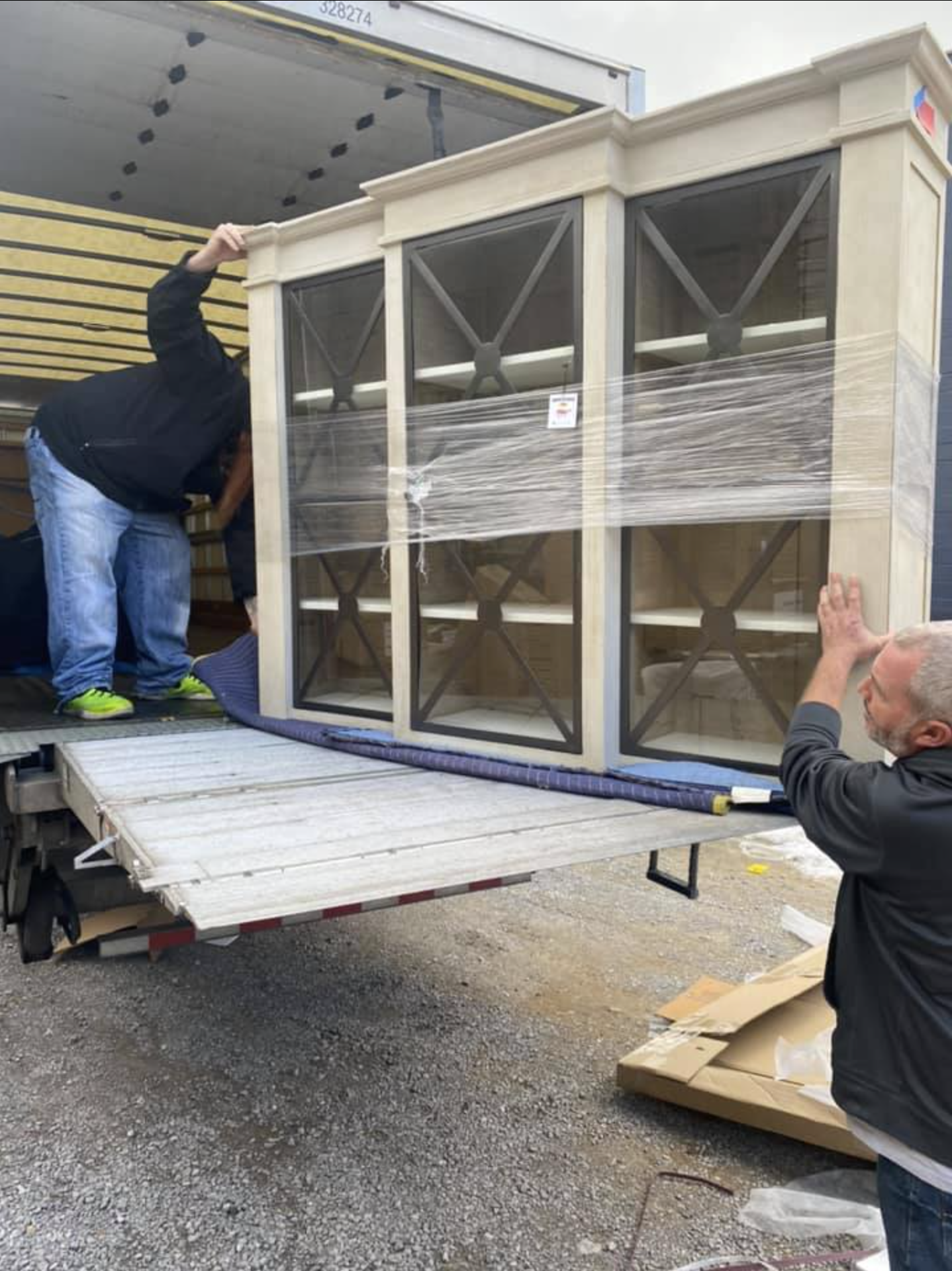 Two men are unloading a large, beige wooden cabinet with glass doors from a truck. One man is standing on the truck, the other on the ground, both guiding the piece. The cabinet is wrapped with plastic for protection.