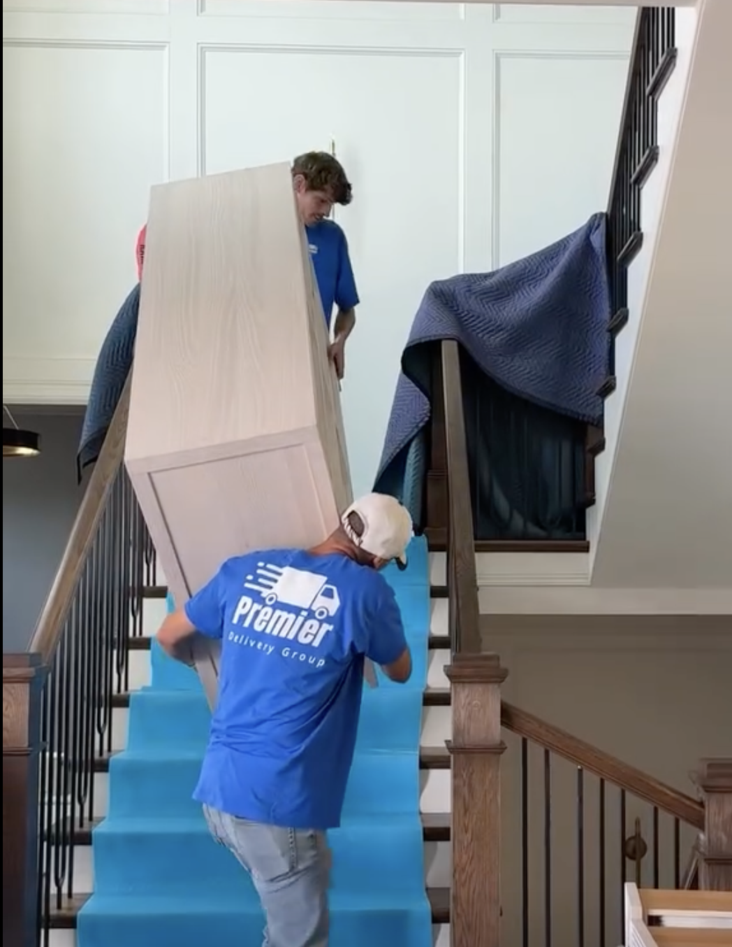 Two movers loading a large wooden cabinet up a staircase, with one standing on the upper level and the other on the stairs, wearing a blue Premier Delivery Group shirt.