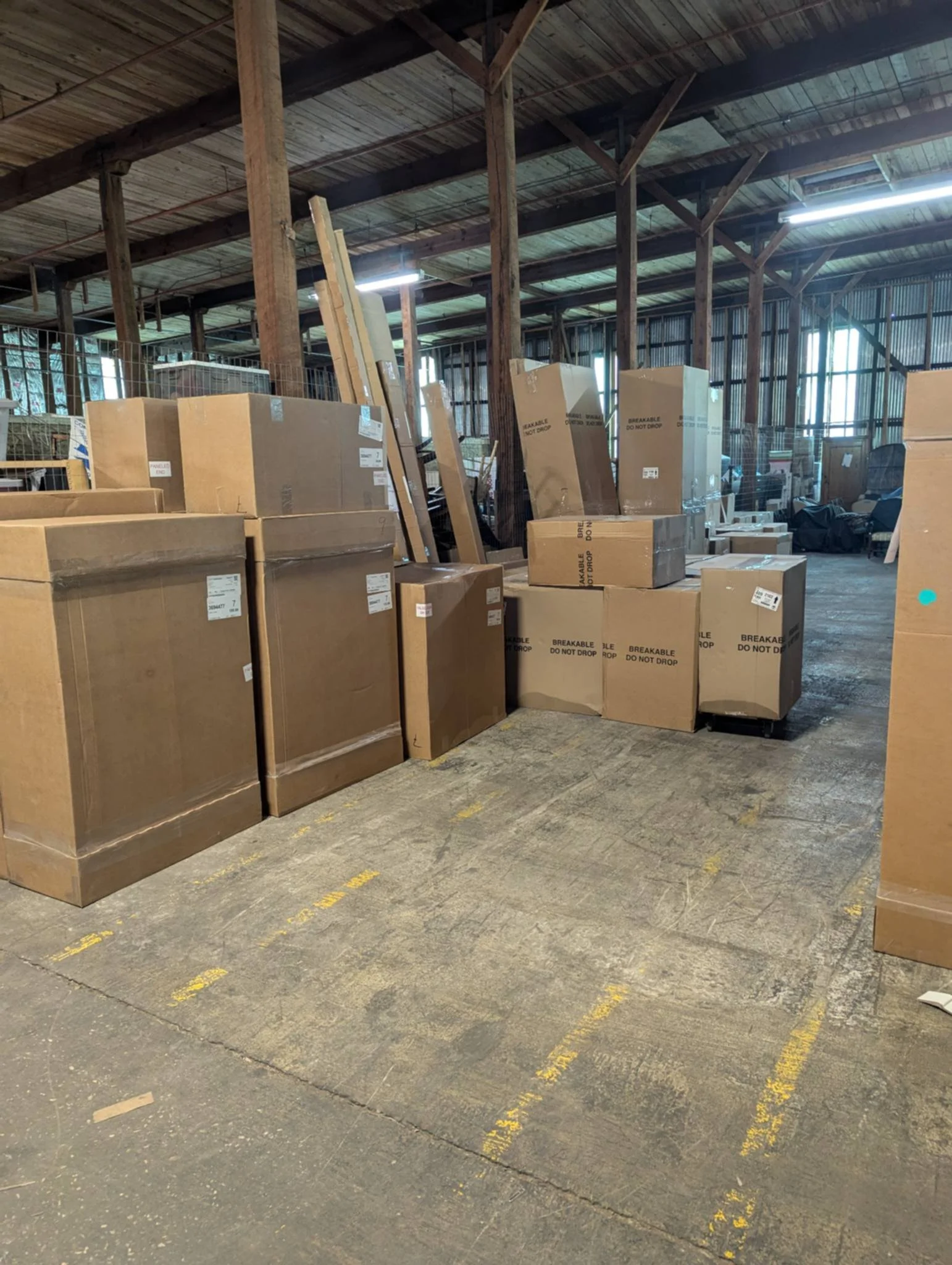 Storage area with numerous large cardboard boxes and wooden planks inside a warehouse with wooden beams and metal walls.