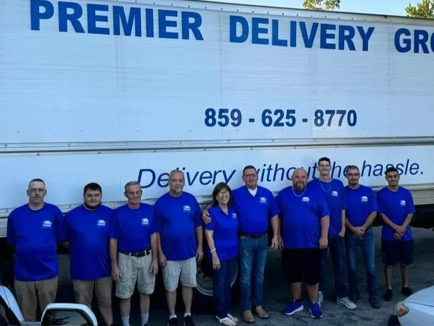 Group of ten people standing in front of a delivery truck, all wearing matching blue shirts with a logo, smiling for the photo.