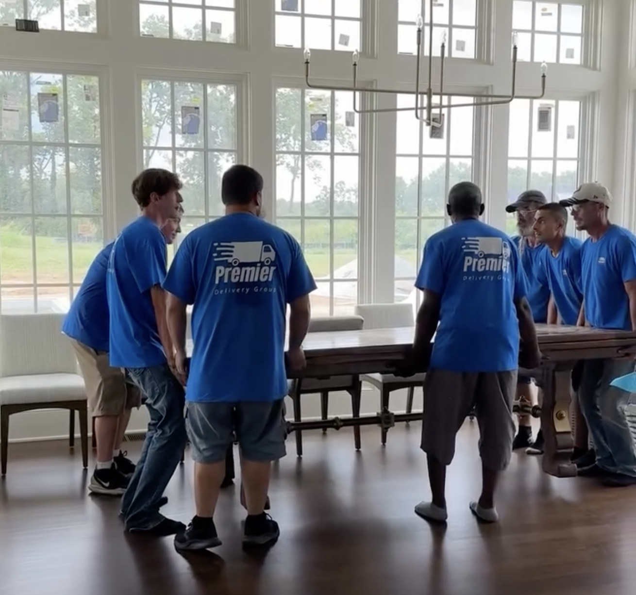 A group of people wearing blue 'Premier Delivery Group' shirts working together to move a long piece of wood furniture in a bright room with large windows.