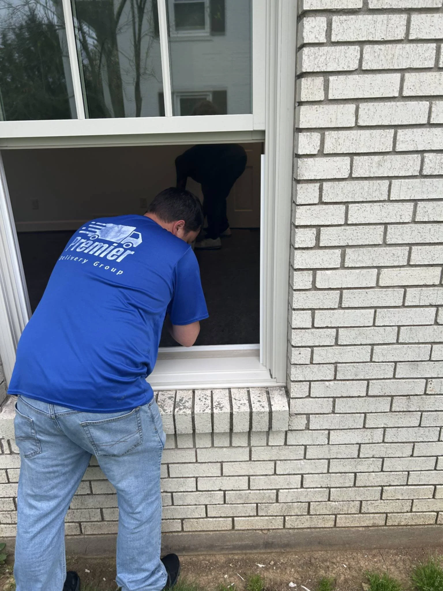 A worker in a blue Premier delivery group shirt installing or repairing a window on a brick house, with another worker inside the house visible through the window.