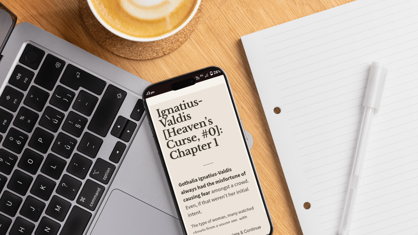 A workspace with a laptop, a smartphone displaying the start of a book titled "Heaven's Curse" by Ignatius-Valdis, a cup of coffee, a blank notepad, and a pen on a wooden desk.