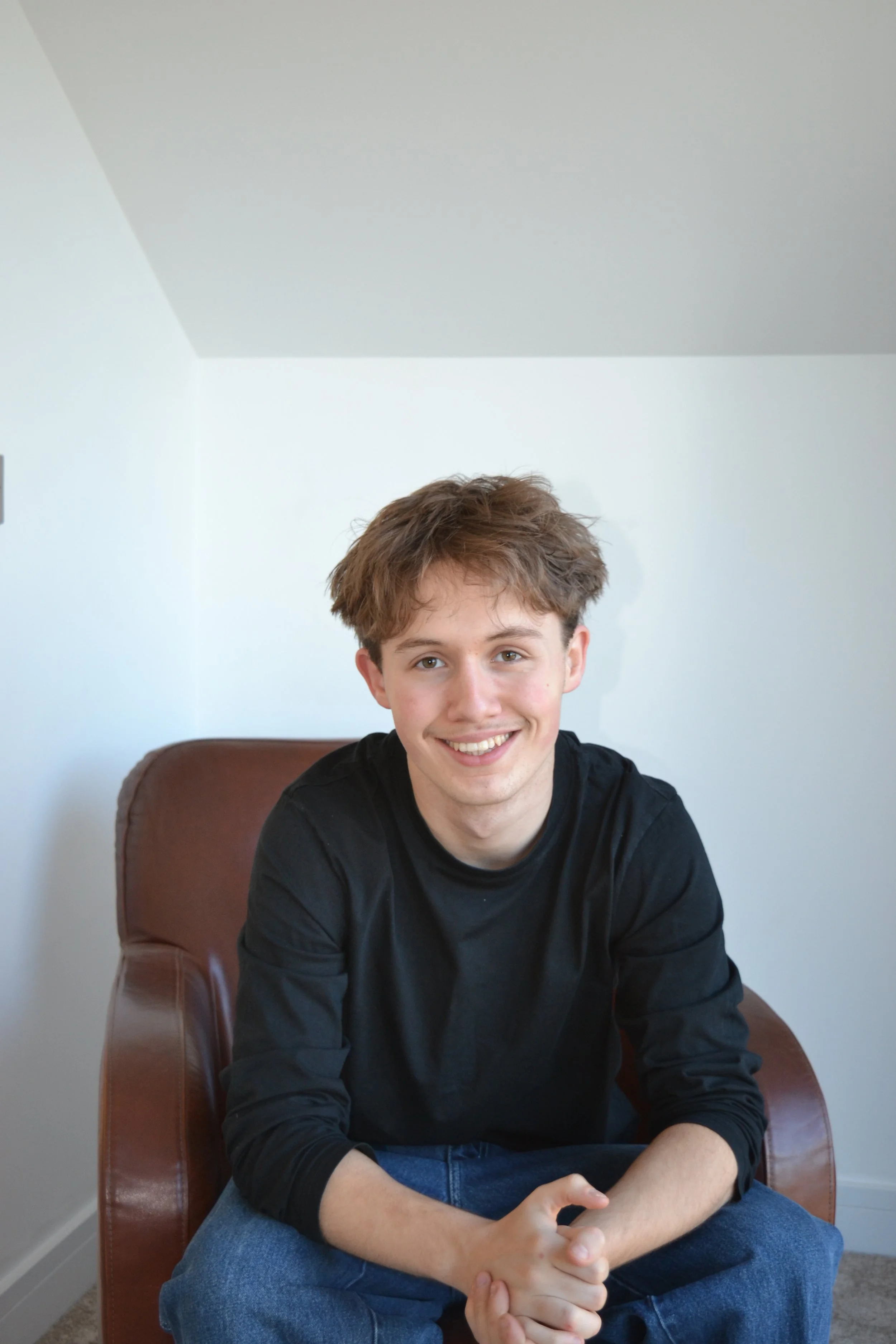 A young man with brown, tousled hair smiling while sitting on a brown armchair against a plain white wall.