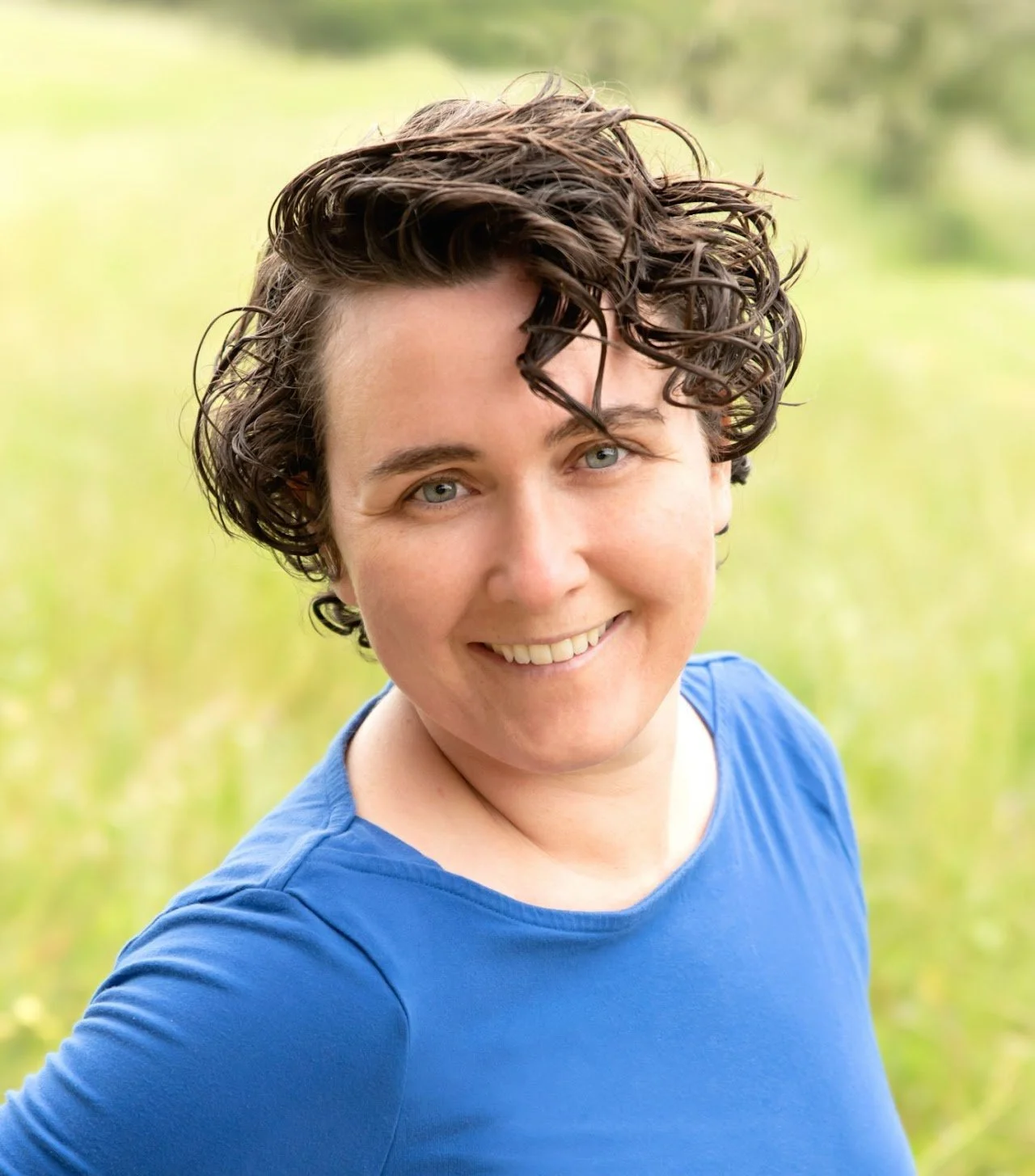 A woman with short, curly brown hair and blue eyes smiling outdoors, wearing a blue shirt with a blurred green background.