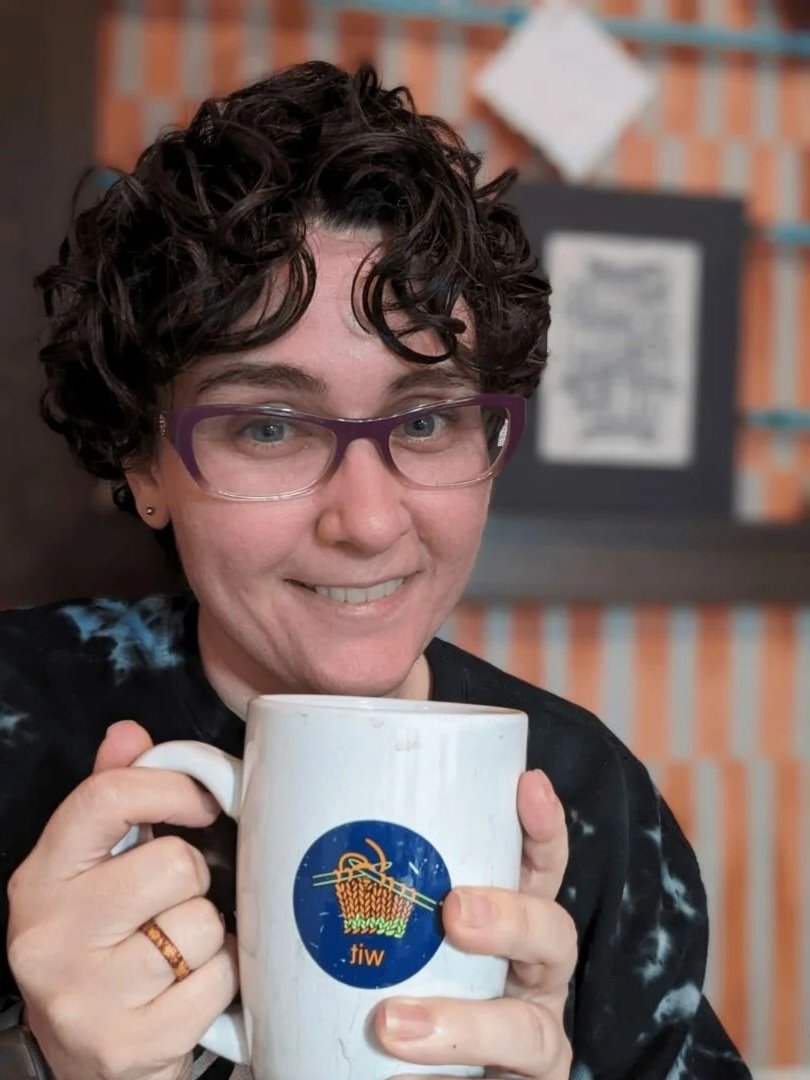 Woman with short curly hair, purple glasses, and a ring on her finger holding a white mug with a knitting logo and the word "witi" in front of a brick wall