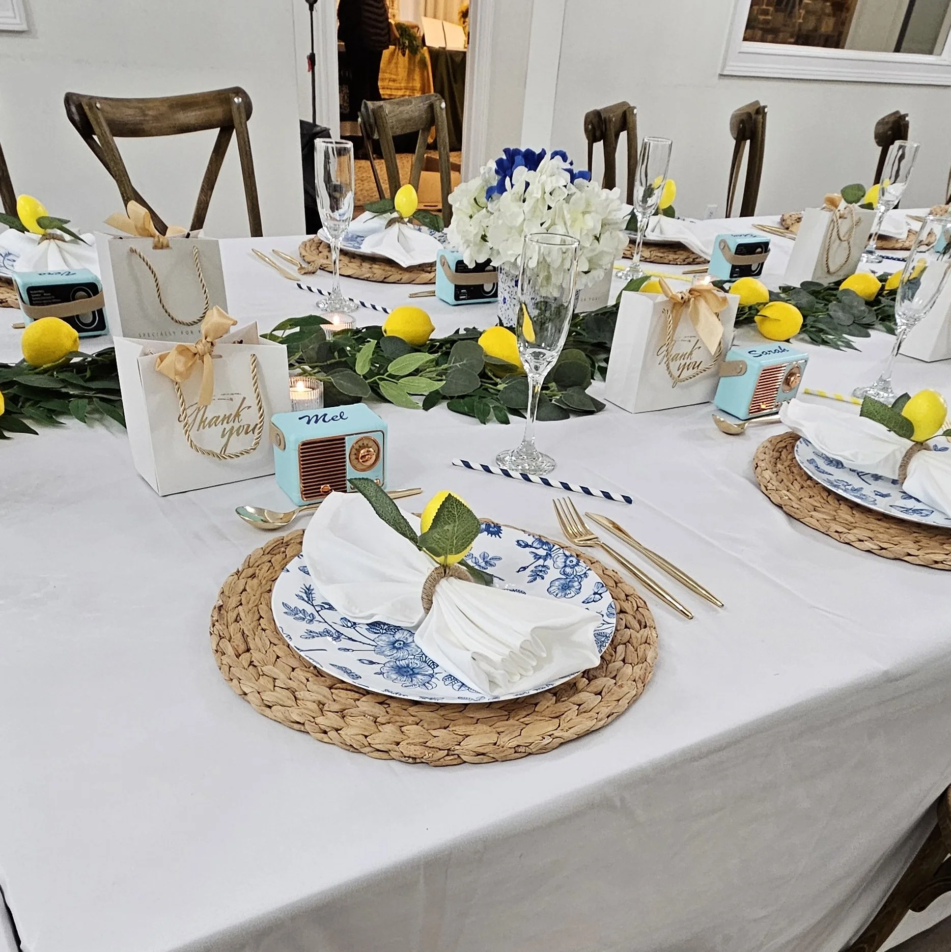 A decorated dining table with blue and white floral plates on woven placemats, white napkins tied with green leaves and small yellow lemons, gold utensils, clear champagne glasses, and small blue radios labeled 'Me' and 'Thank You'. A green garland with yellow lemons runs along the center, with a floral centerpiece of white and blue flowers. Gift bags with gold lettering are also on the table, and a large window is visible in the background.