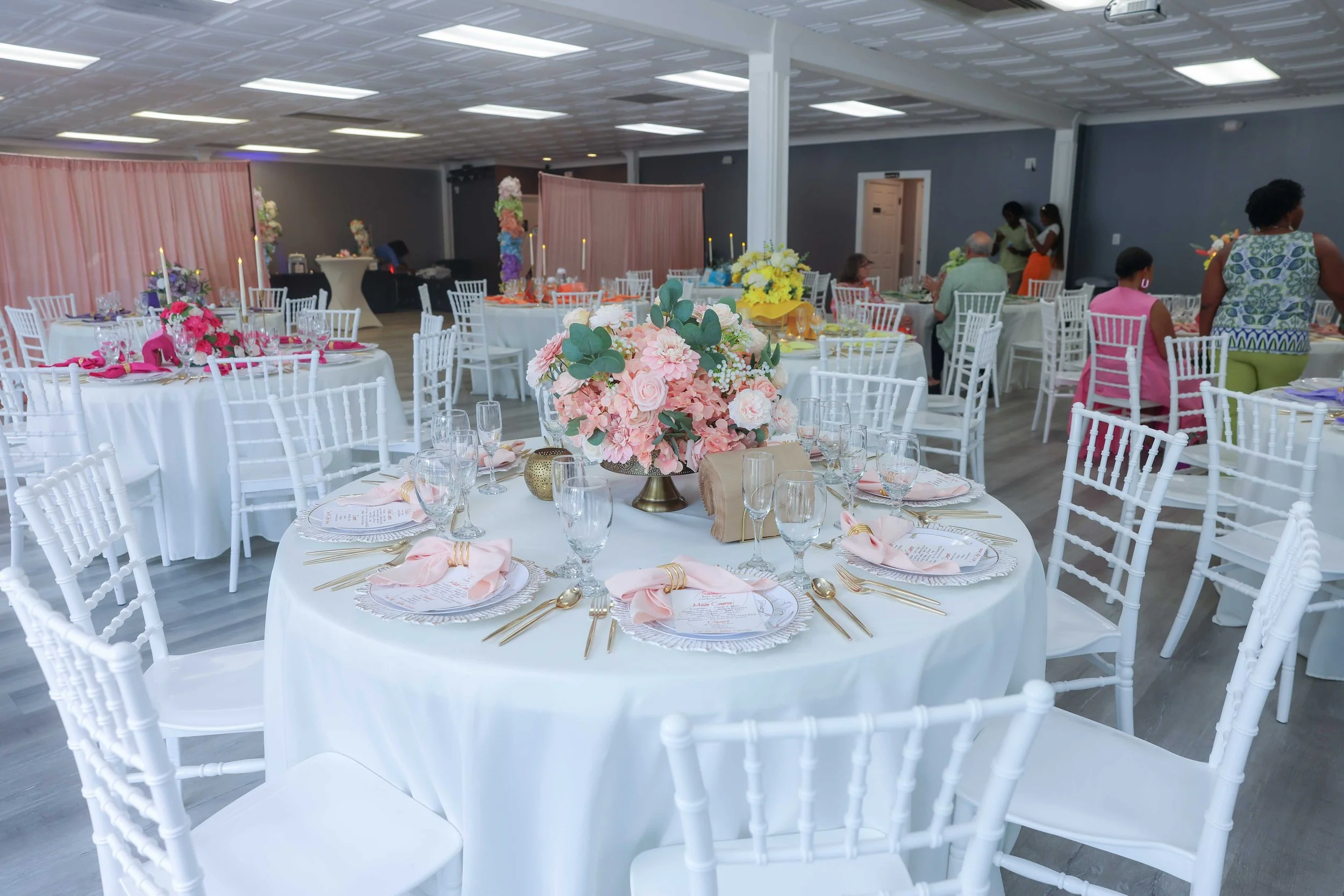 Event space decorated with round tables covered in white tablecloths, featuring pink and floral centerpieces, set for a celebration or wedding reception.