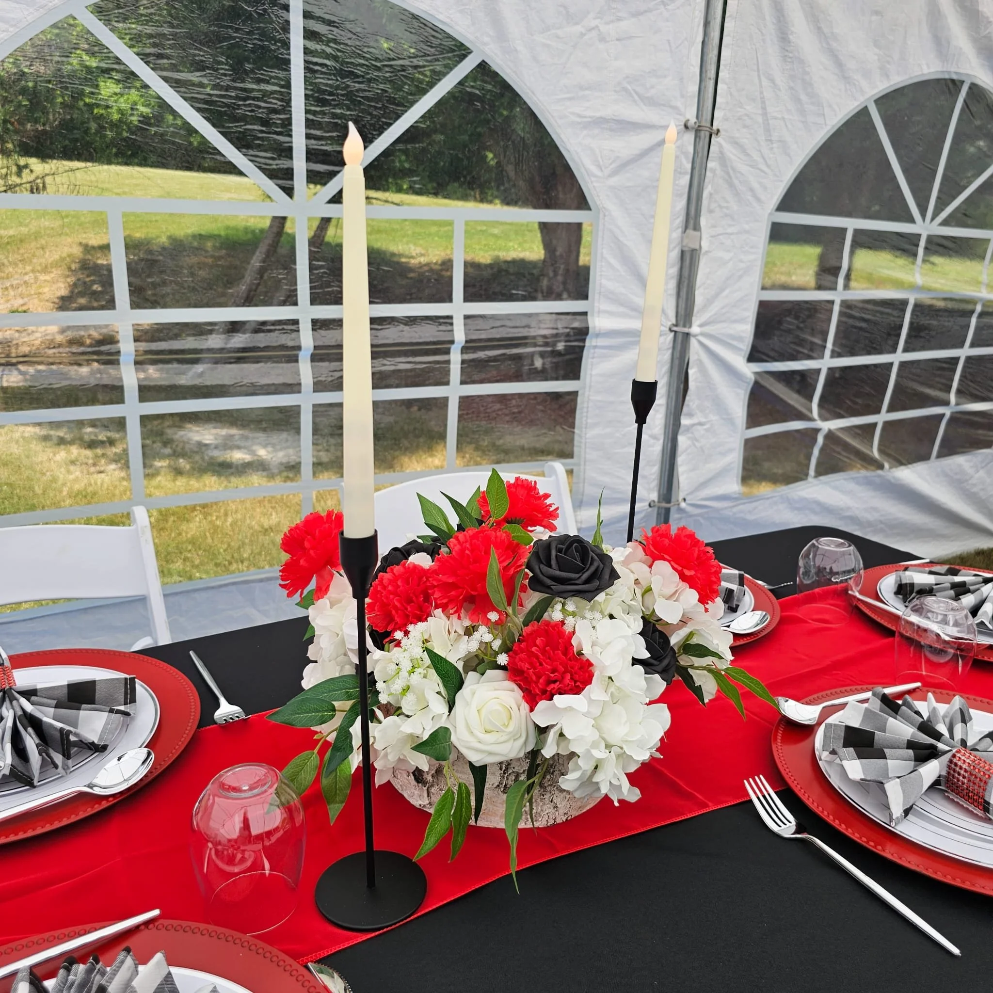 A decorated outdoor table with a black tablecloth, red table runner, floral centerpiece with red, black, and white flowers, candles, wine glasses, and black and white checkered napkins inside a white tent.