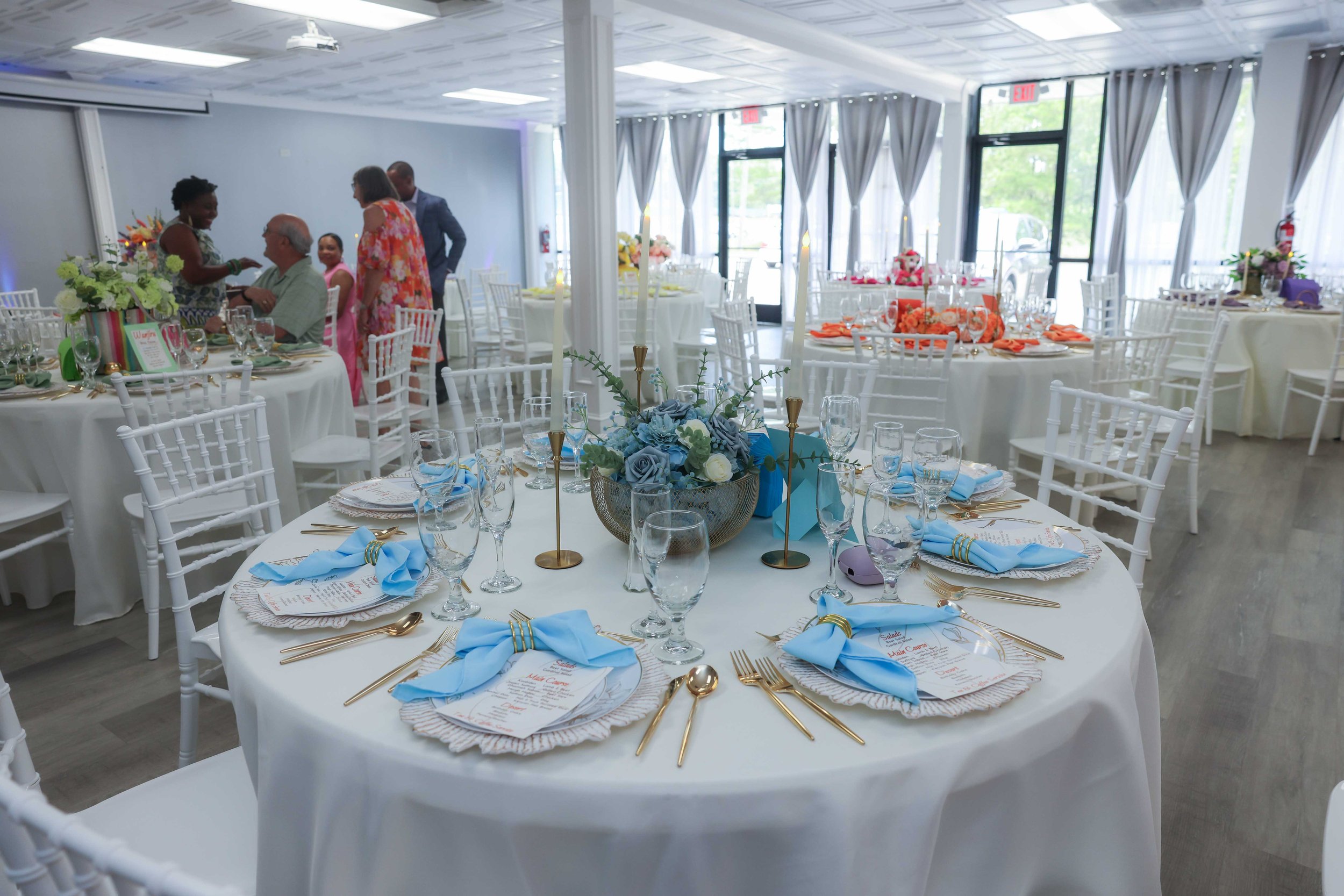 Event space with round tables decorated with blue, orange, and purple floral centerpieces, set for a celebration with place settings, napkins, and gold cutlery. Multiple chairs surround each table, and people are mingling in the background.