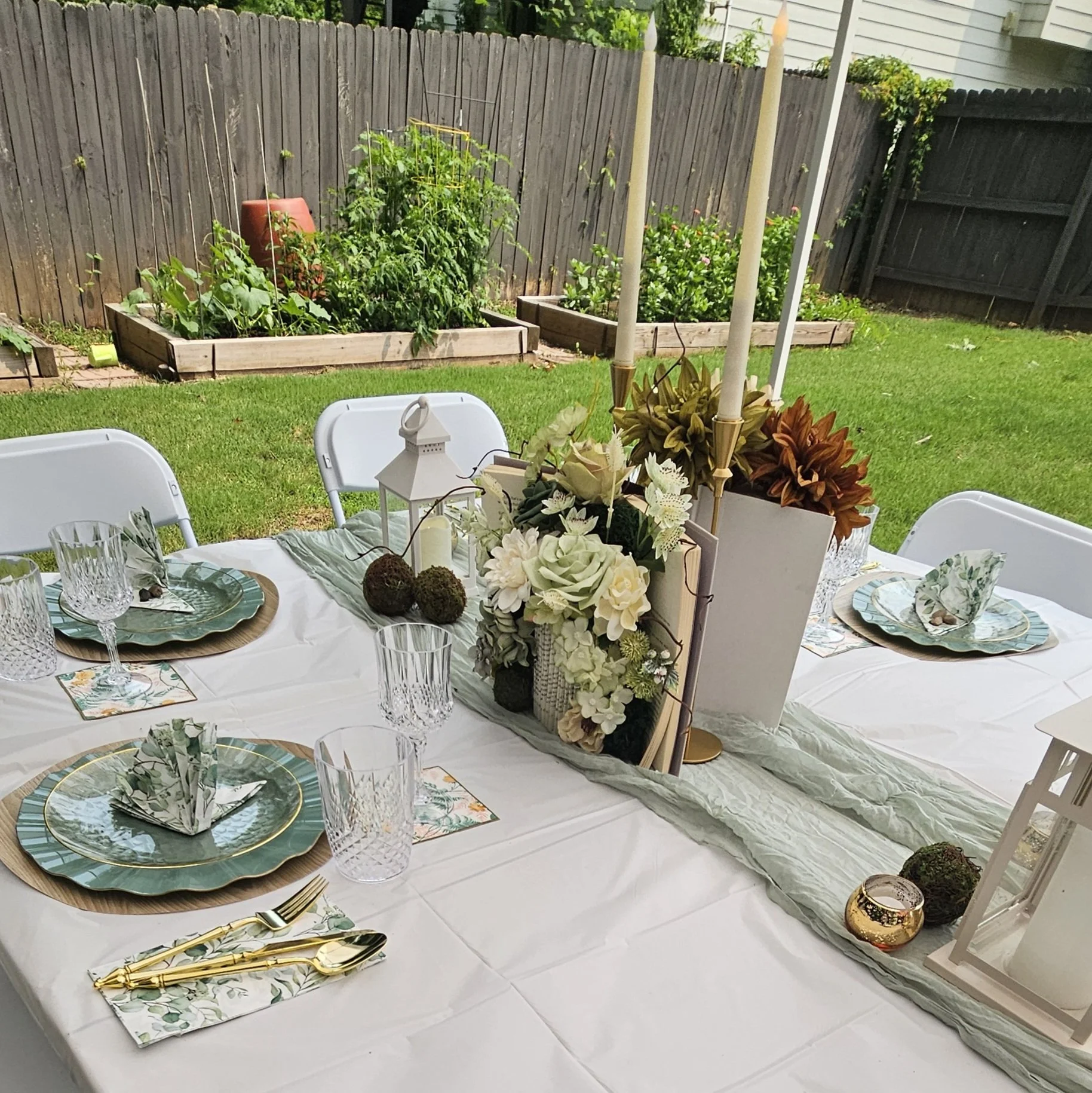 Outdoor dining table decorated with floral arrangements, candles, and gold cutlery, set with clear glassware and patterned plates in a backyard with green grass and garden beds.