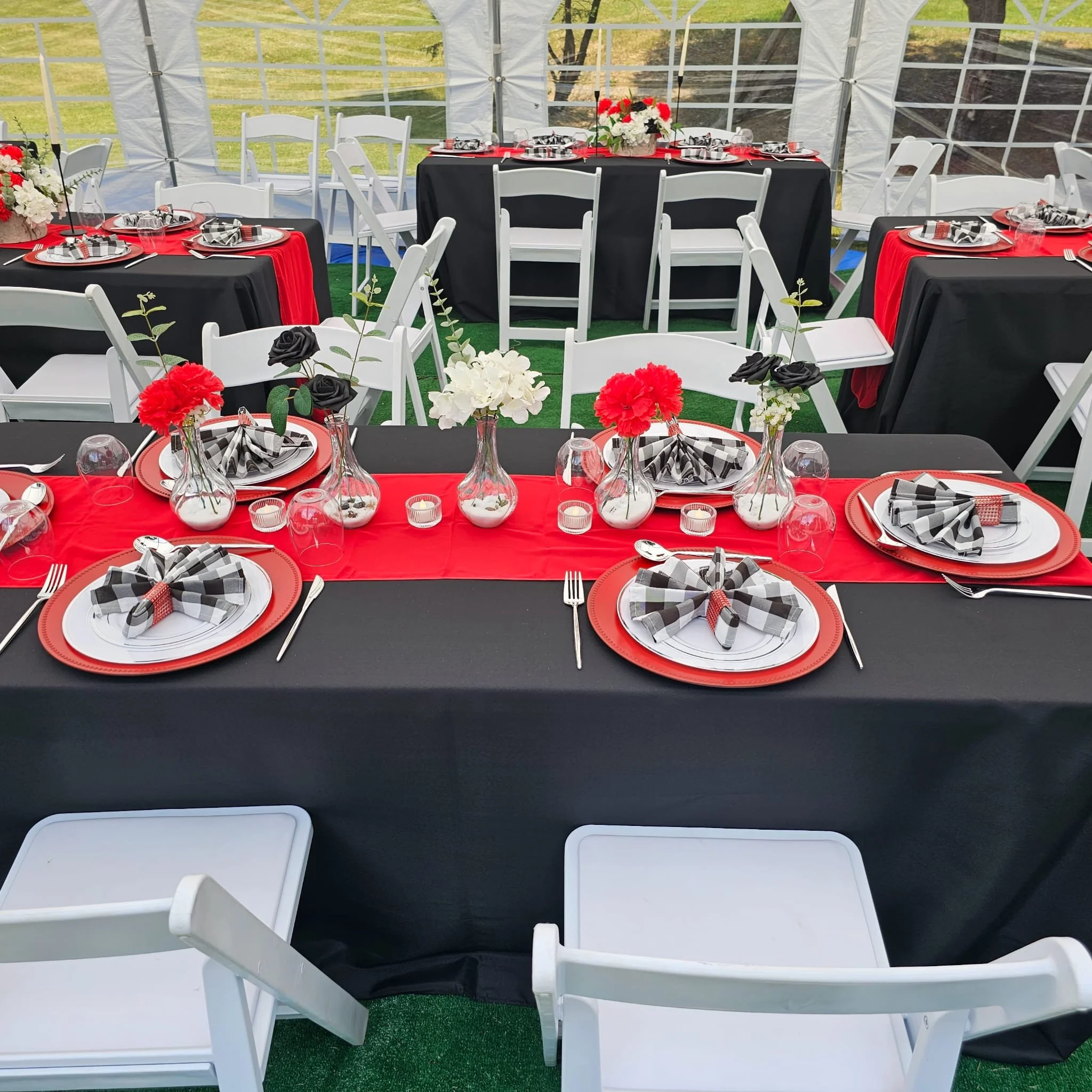 Decorated event tent with tables featuring red and black tablecloths, set with plates, checkered black and white napkins, cutlery, vases with red, black, and white flowers, and small candle holders.