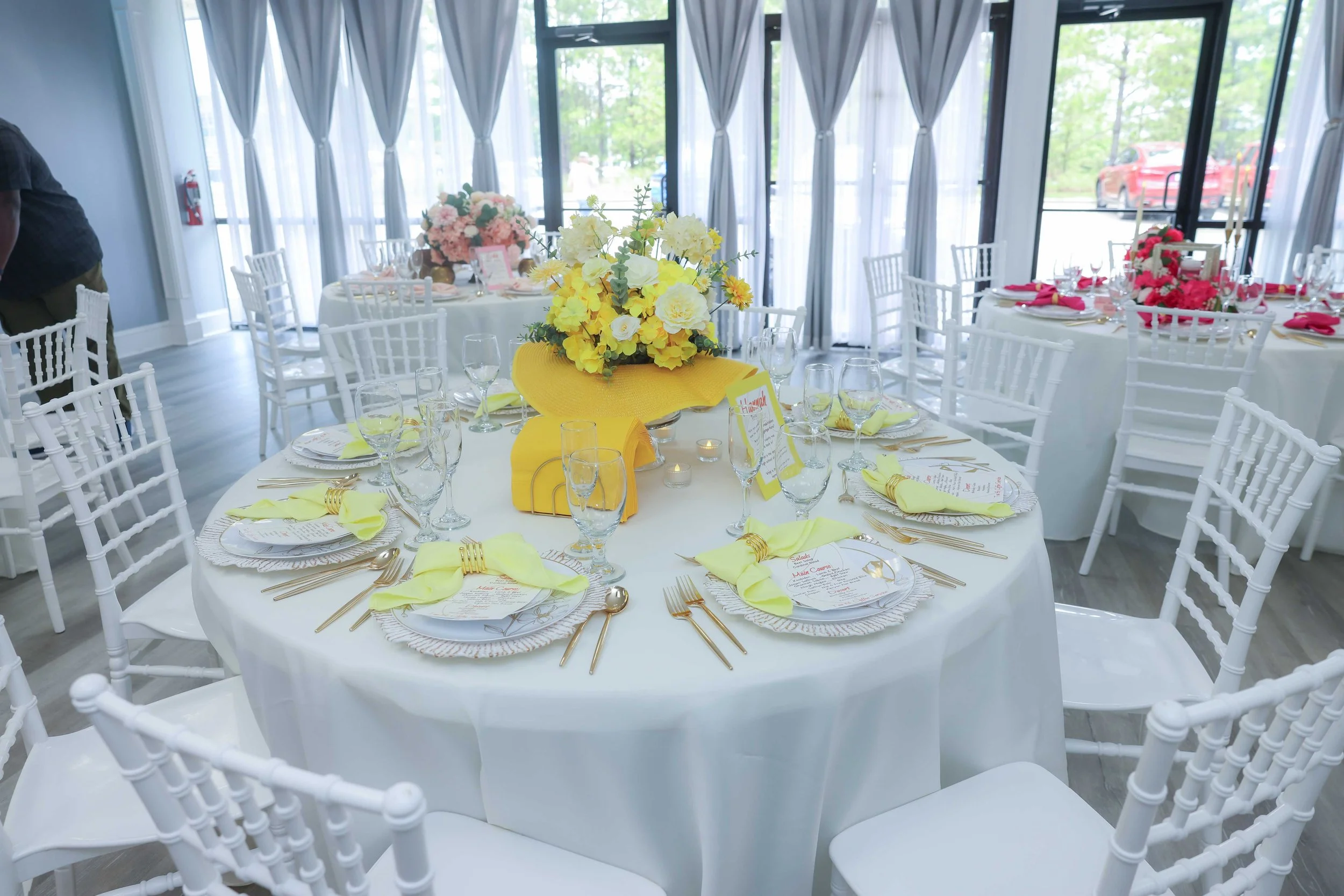 Table set for a formal event with white chairs, yellow and pink floral centerpieces, and yellow napkins in a bright room with large windows.
