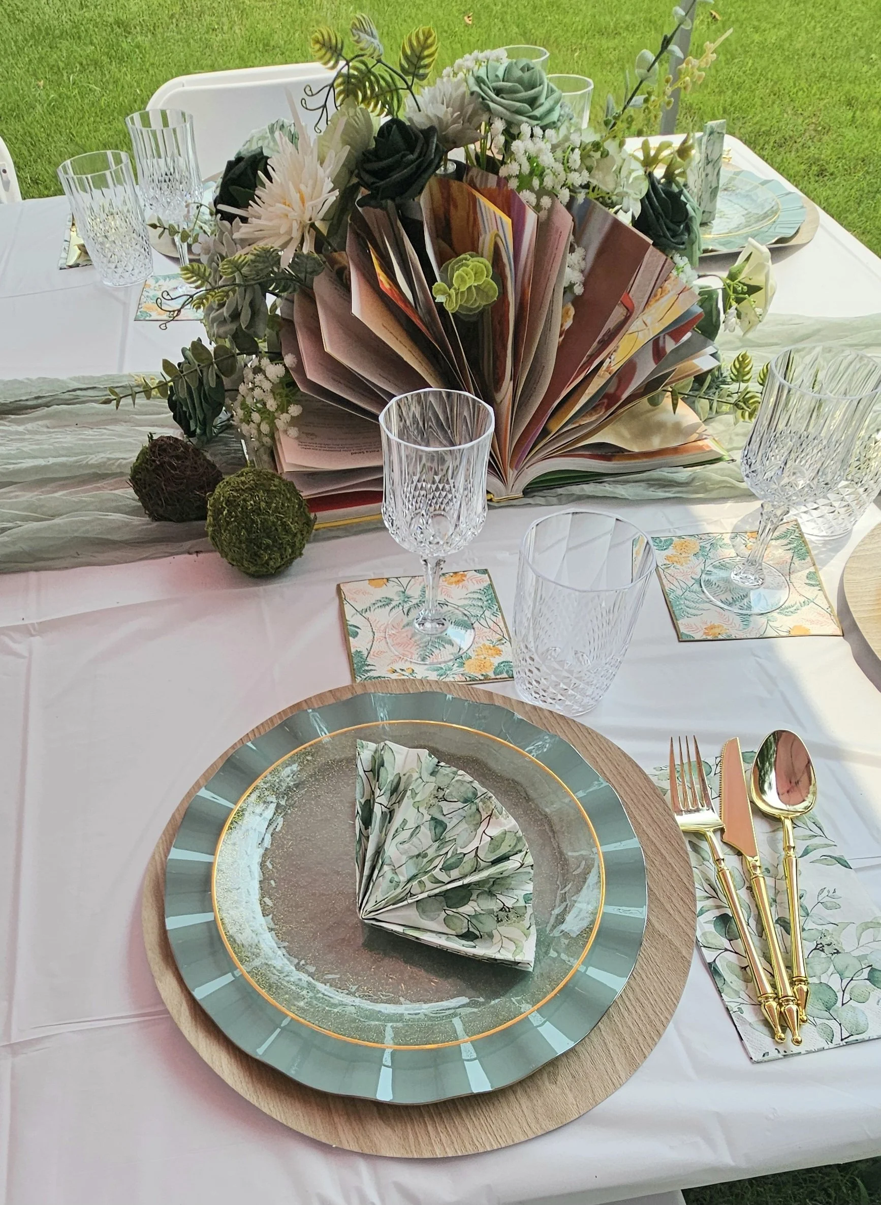 Elegant outdoor dining table decorated with a floral centerpiece, glassware, and place settings, featuring a green and gold theme with folded napkins.