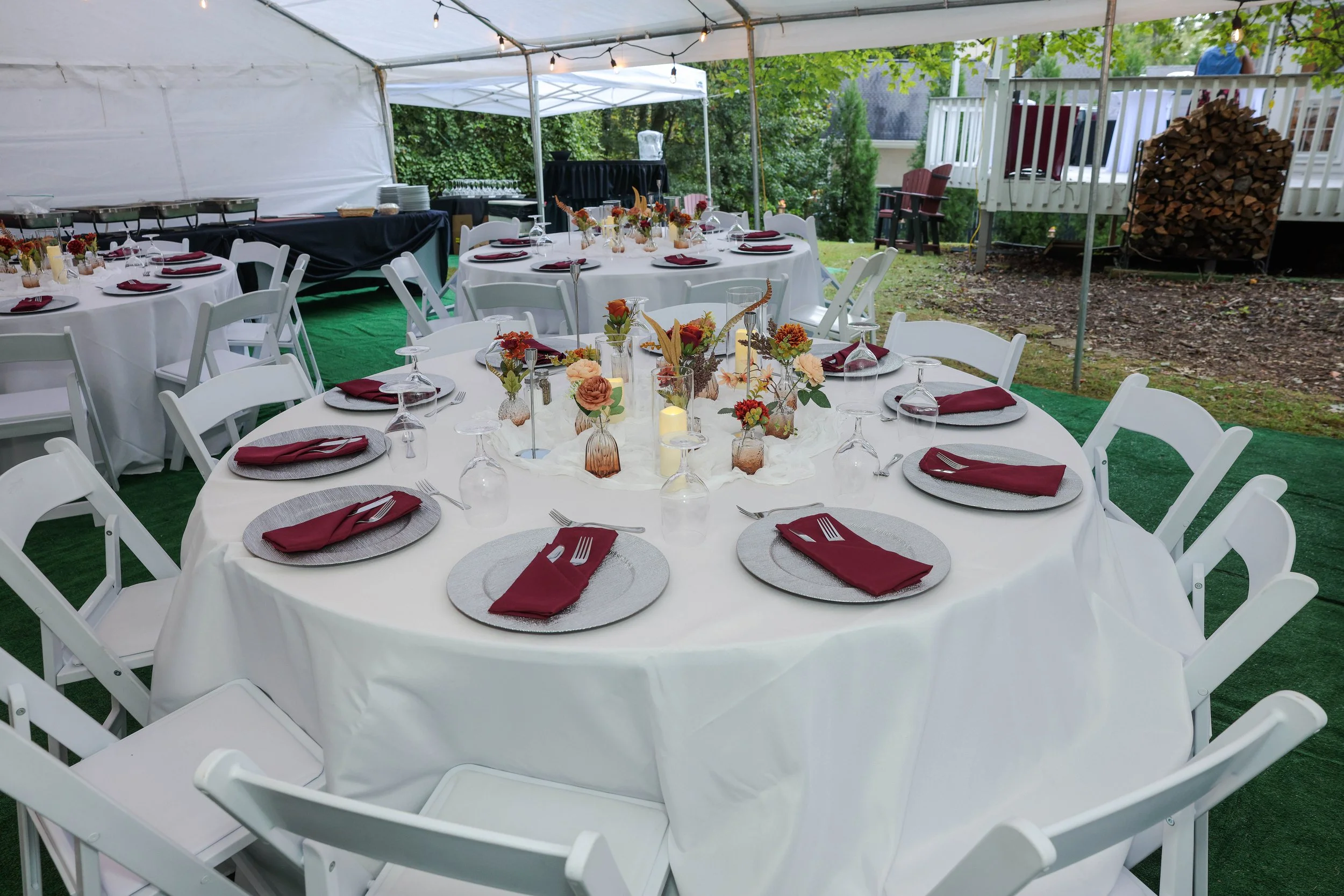 Round outdoor banquet table set with white tablecloth, maroon napkins, and glassware, decorated with autumn-themed floral centerpieces and candles under a white canopy tent.