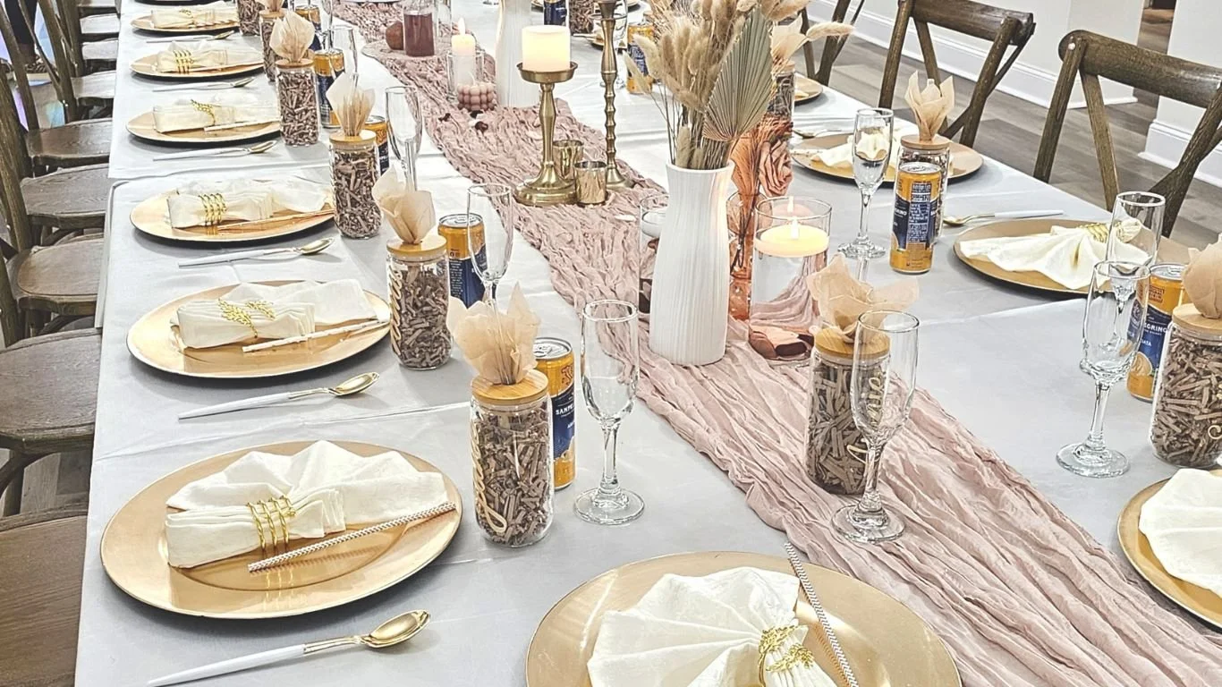 A long dining table set for a celebration with gold and neutral tones, featuring gold plates, white napkins, gold cutlery, glassware, and decorative vases with dried flowers and candles on a pink table runner.