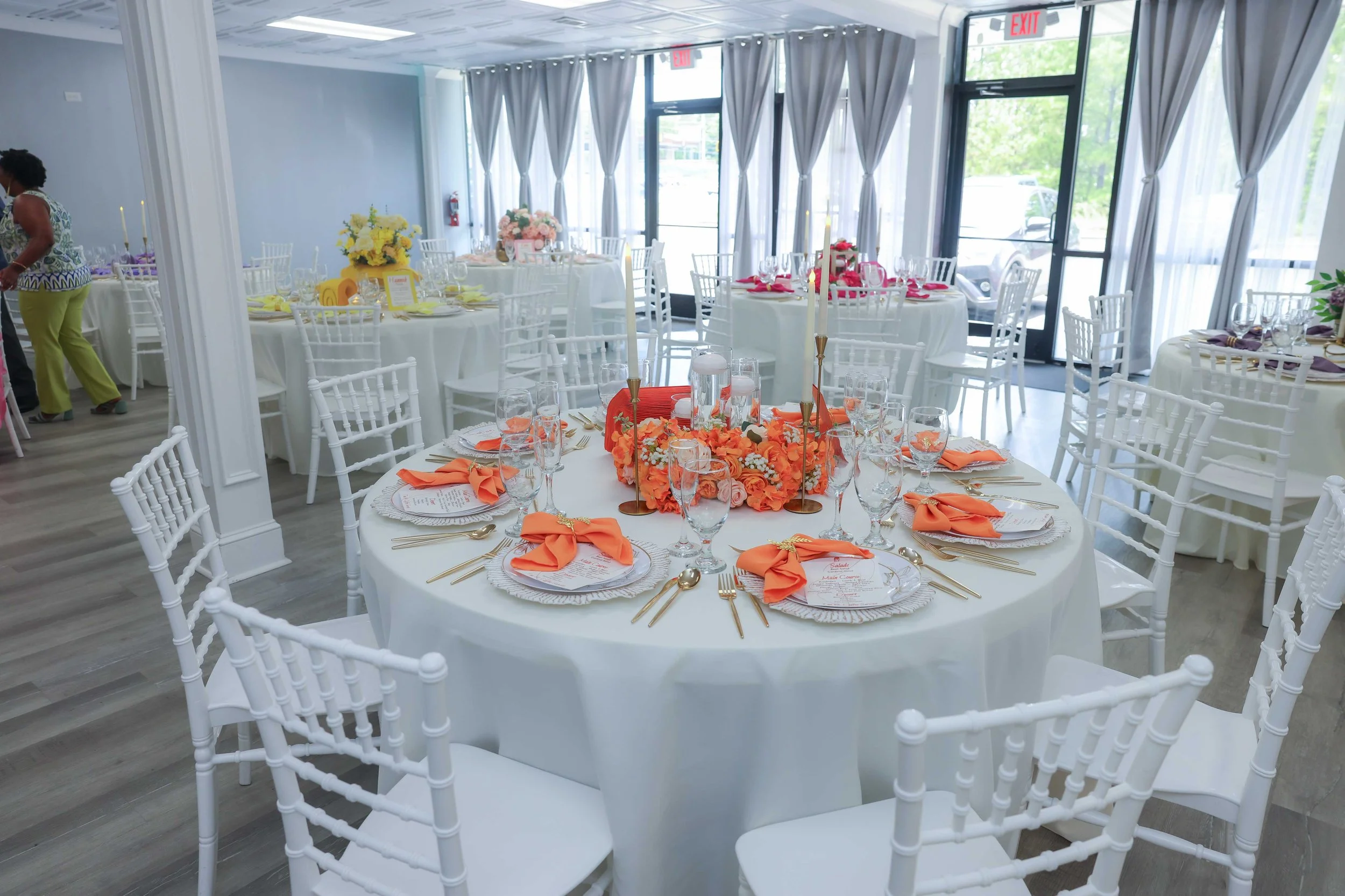 Round table at an event decorated with orange flowers, candles, and orange napkins, set with glassware, gold cutlery, and printed menus in a bright, airy room with large windows and white curtains.