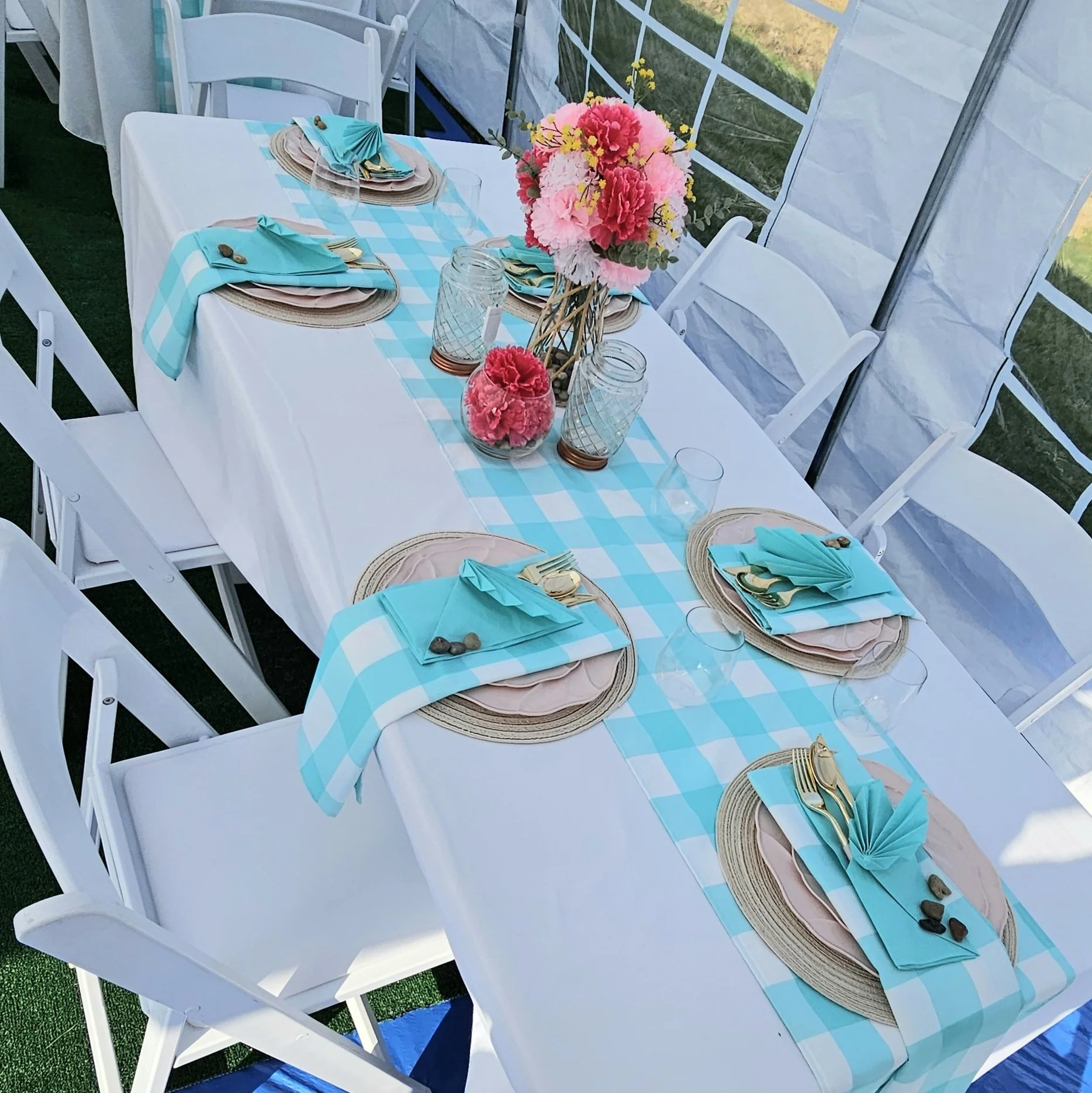 Outdoor table decorated with a blue and white checkered table runner, pink floral centerpiece, and place settings with woven placemats, light pink napkins, and teal napkins folded over the plates, surrounded by white folding chairs.