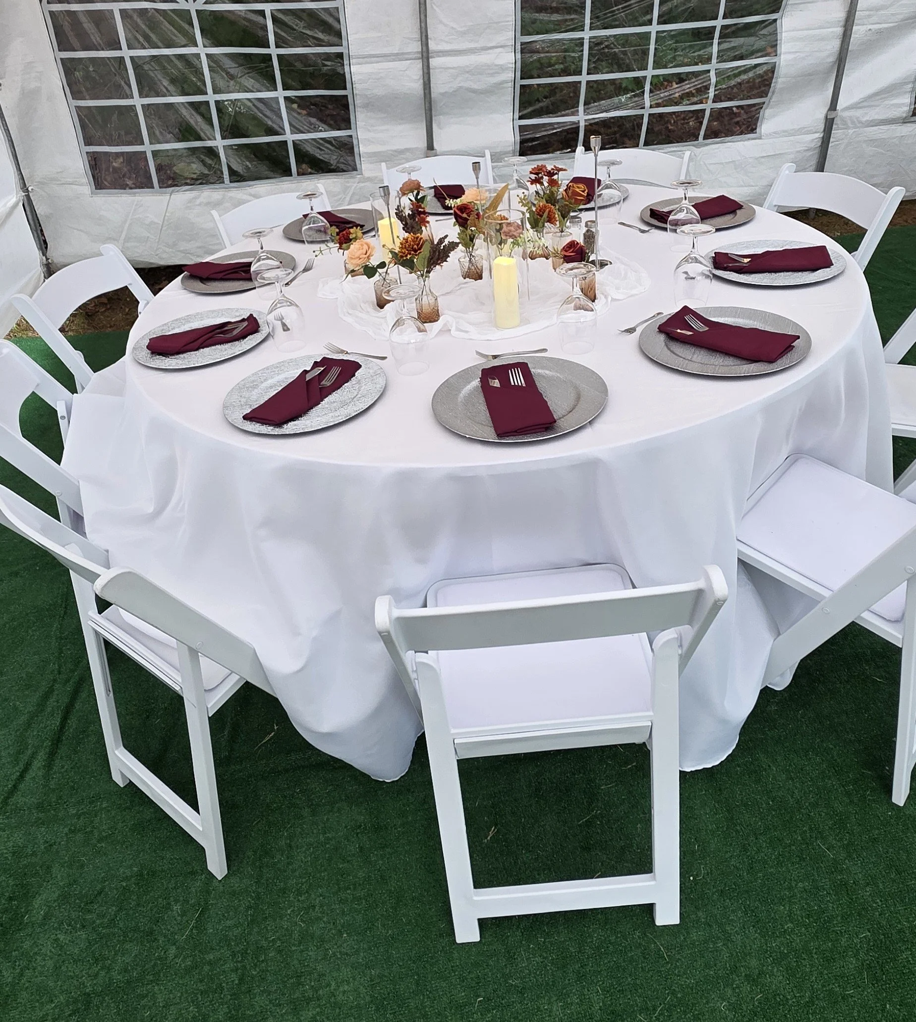 A round table set for a formal event with a white tablecloth, six white chairs, and a centerpiece of flowers, candles, and wine glasses. Each place setting has a silver plate, a maroon napkin, and utensils.