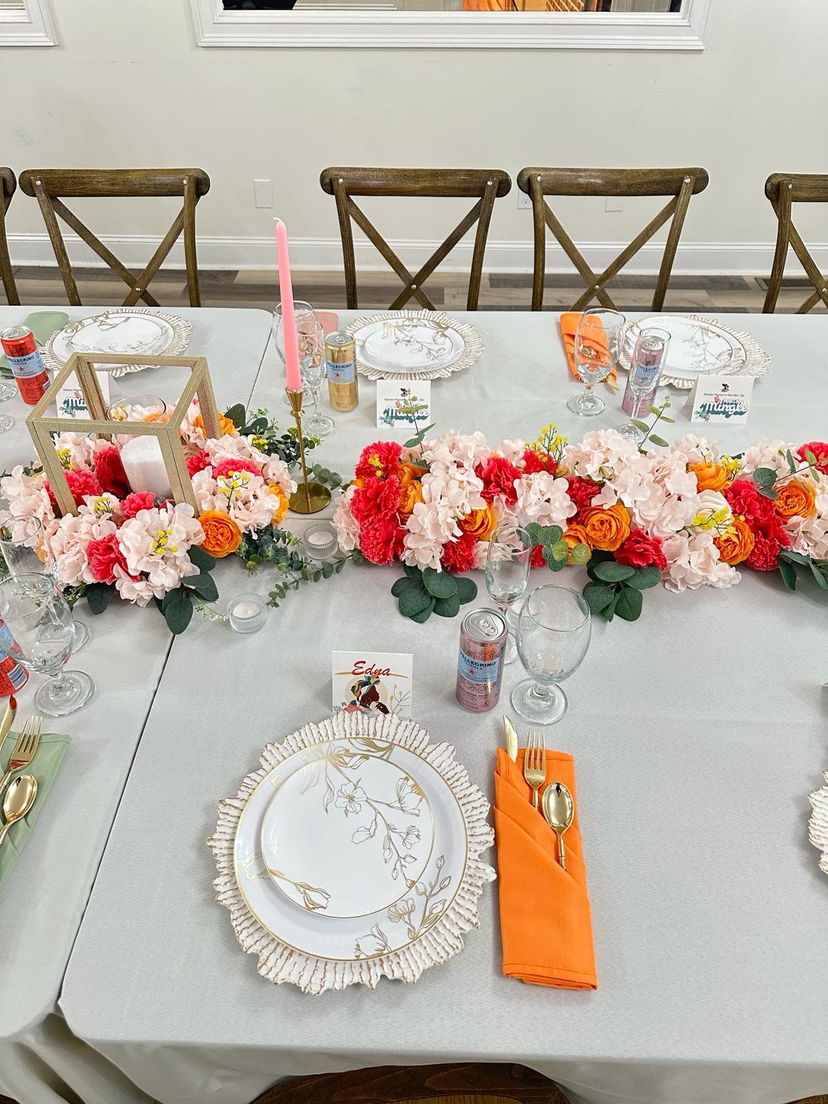 A decorated dining table with floral centerpieces, gold-trimmed plates, orange napkins, gold utensils, and drinks, set for a celebration or gathering.