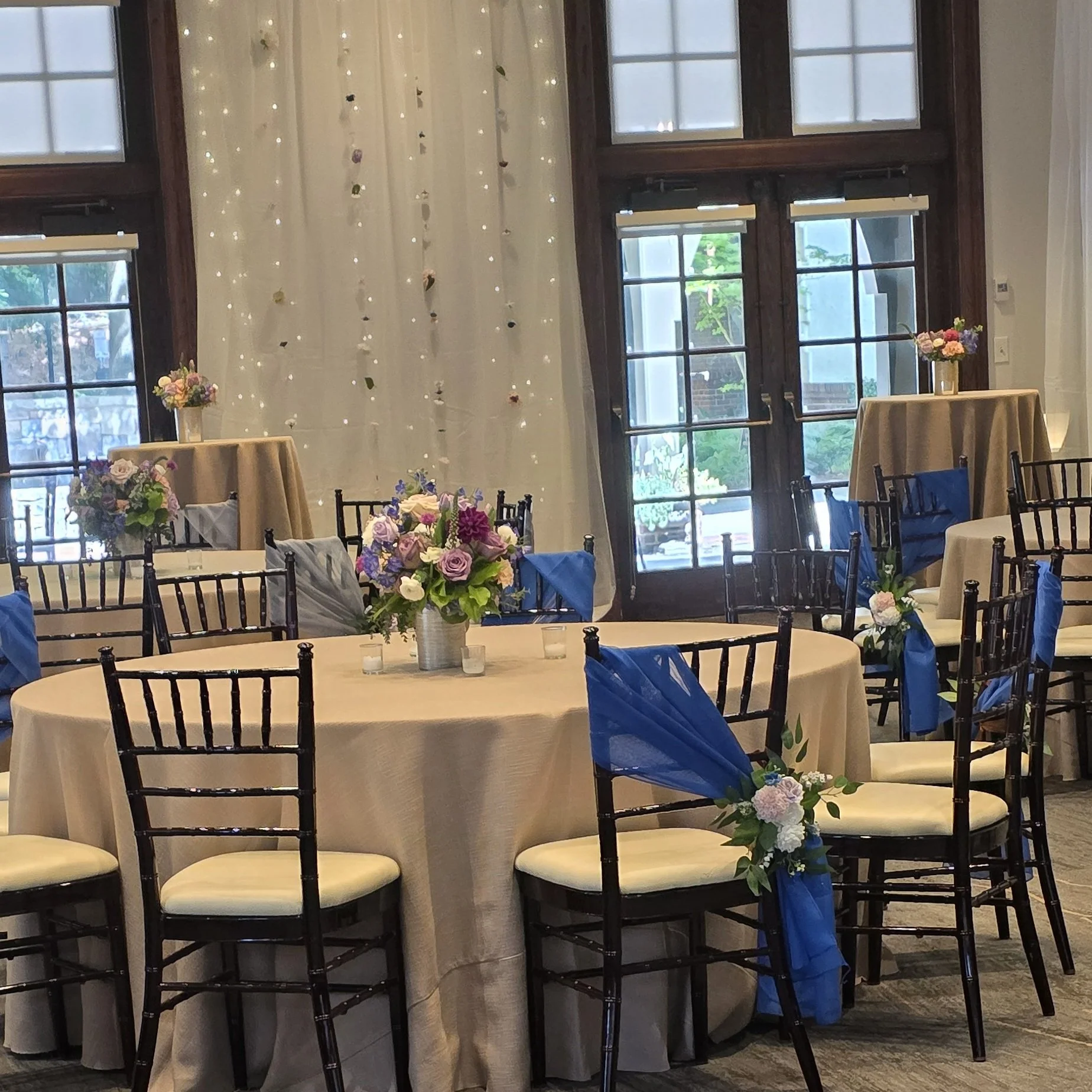 Elegant event space with round tables decorated with beige tablecloths, floral centerpieces, and blue fabric accents on chairs. Tall floral arrangements on high tables and large windows with white curtains and string lights in the background.
