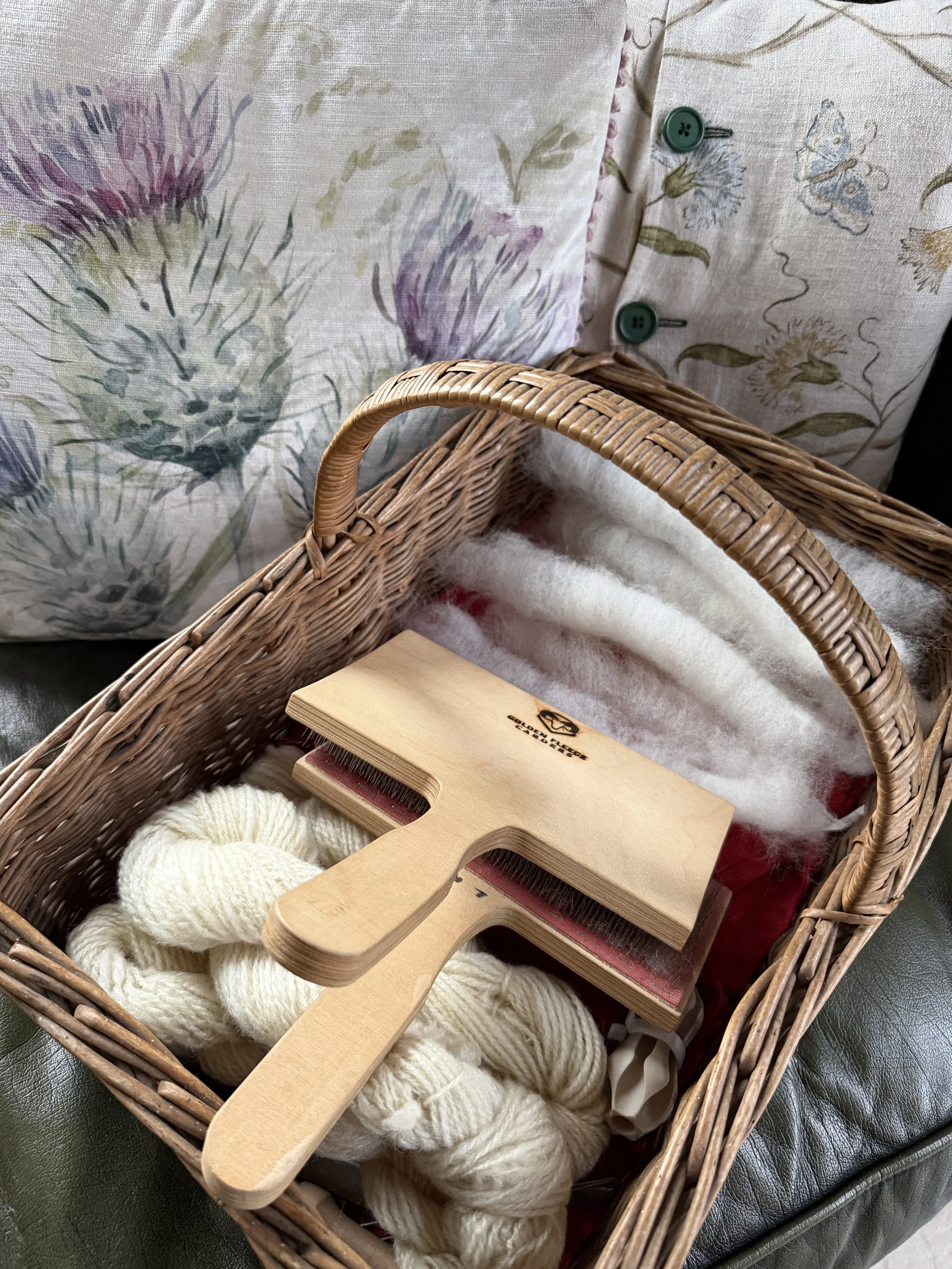 A wicker basket containing skeins of white yarn, some hand carders, and some soft and airy rolags ready to spin, placed in front of floral pillows with button details.