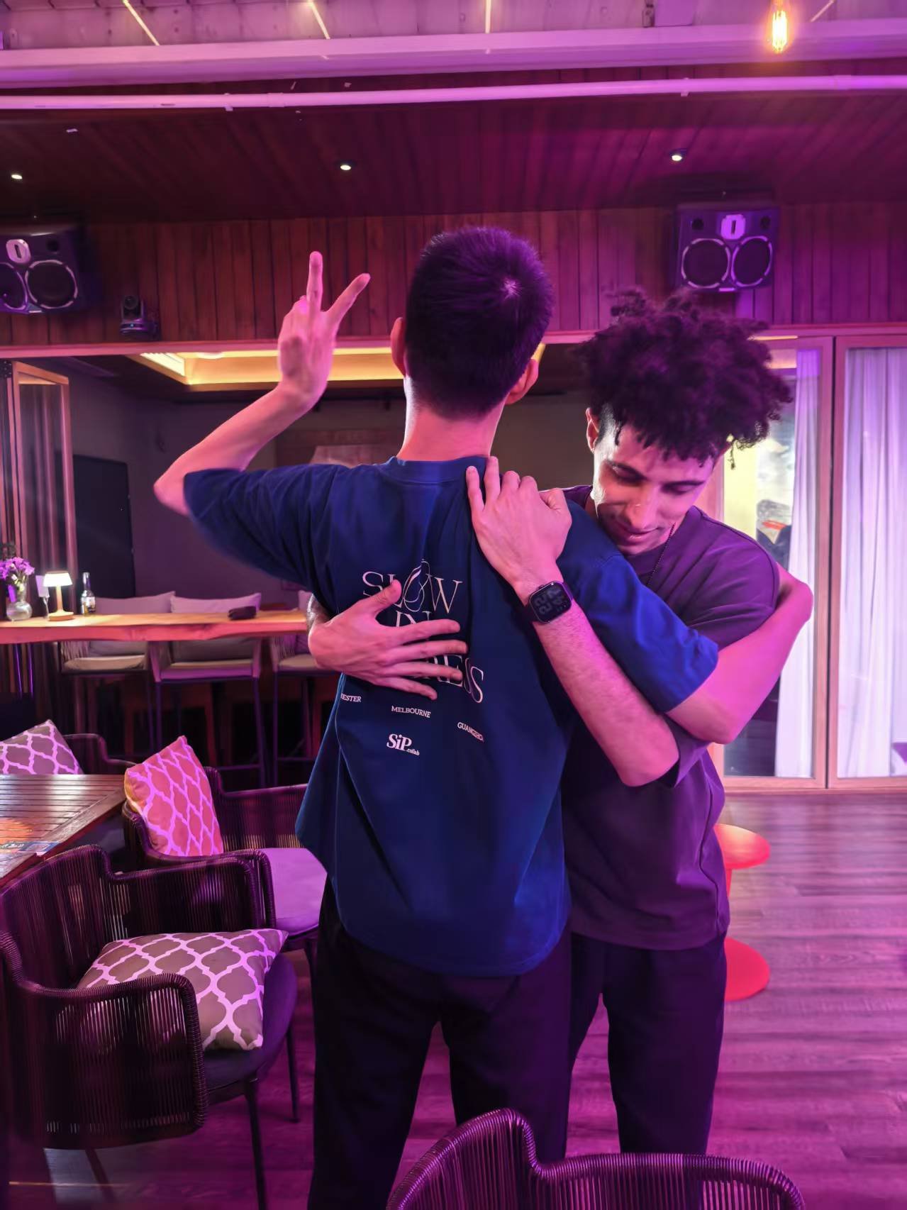 Two men embrace in a hug, one making a peace sign with his fingers, in a dimly lit indoor setting with purple lighting, wooden walls, chairs with patterned cushions, and a bar area in the background.
