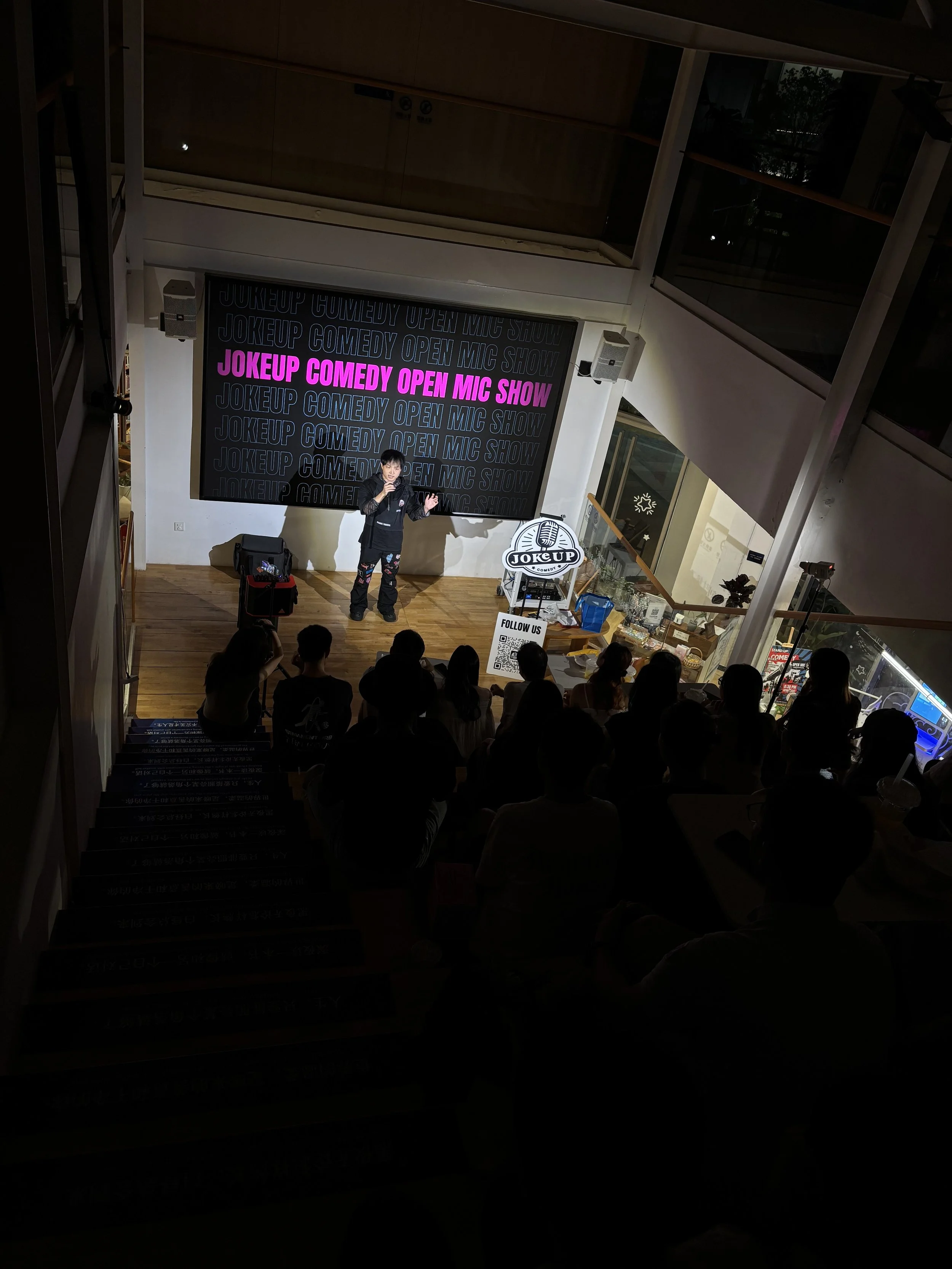 A comedian performing on stage at the Joke Up Comedy Open Mic Show, with a large screen behind displaying the event name and a logo on a stand in front of the stage, audience members seated and watching.