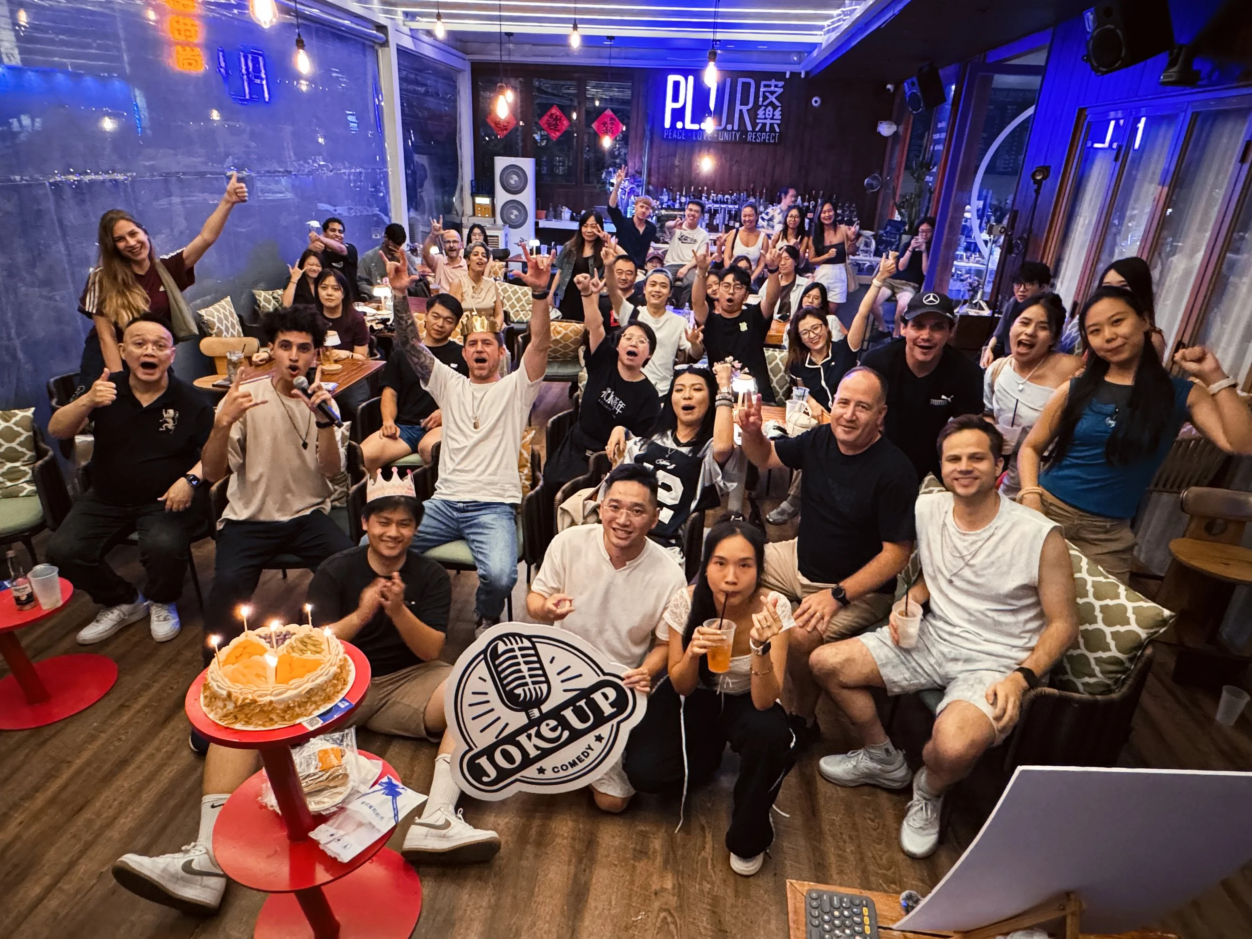 A large group of people gathered in a restaurant or bar, celebrating and smiling. There is a birthday cake with candles in the foreground, and some individuals are making peace signs or raising their hands. The background features a bar area, neon lights, and decorative red hanging ornaments.