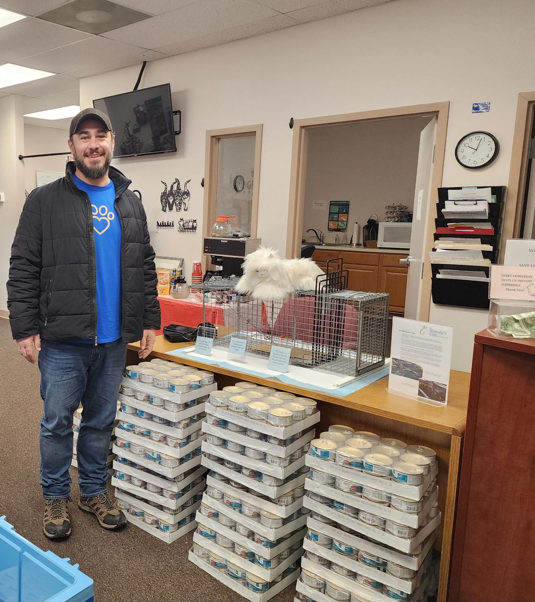 A man with a beard and a cap, wearing a blue shirt and black jacket, standing in front of stacks of canned food inside a room with a TV mounted on the wall and a kitchen area in the background. There are various signs and decorations on the walls, including a clock showing approximately 11:07.
