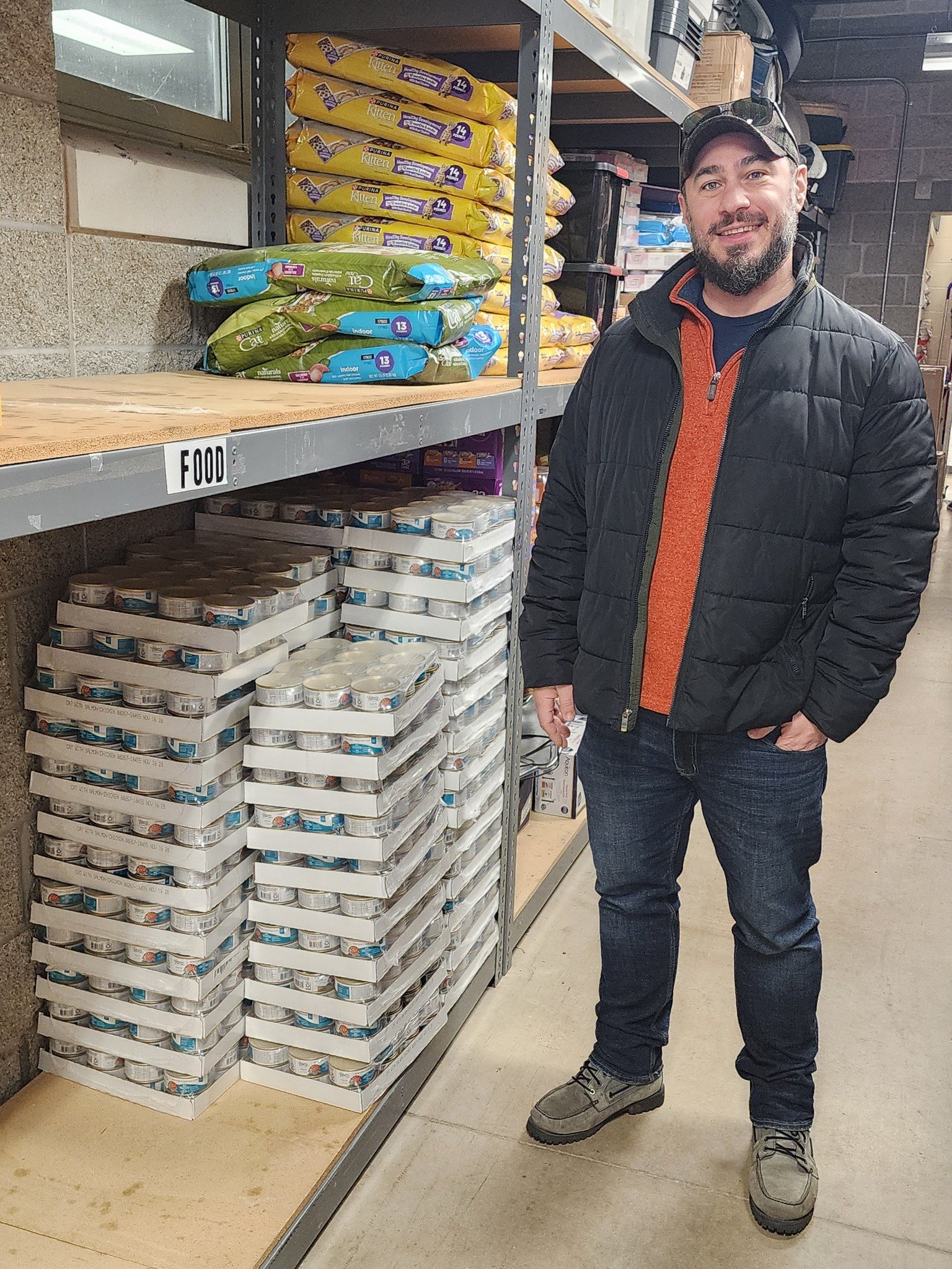 A man wearing a black jacket, orange sweater, jeans, and gray boots standing in an aisle of a store with shelves filled with canned food and pet food. He is smiling and looking at the camera.