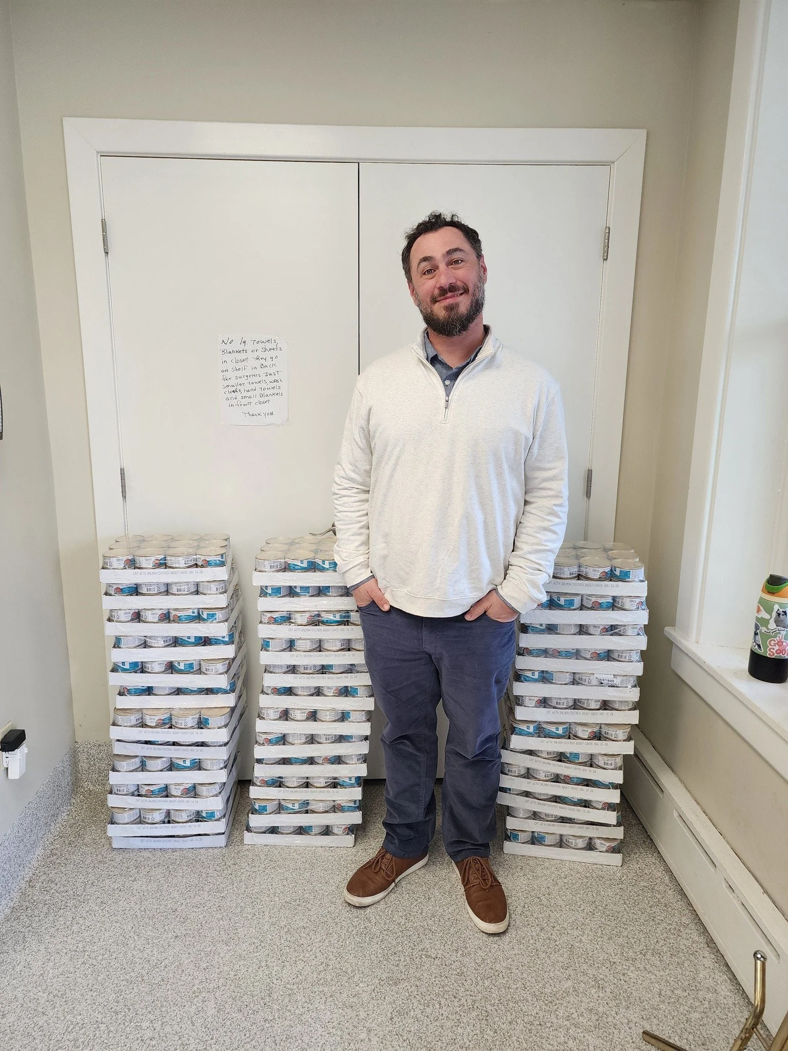 A man with dark hair and a beard stands indoors in front of stacked pallets of canned food, smiling at the camera. The pallets are arranged symmetrically behind him, and there is a wall with a whiteboard and a note in the background.