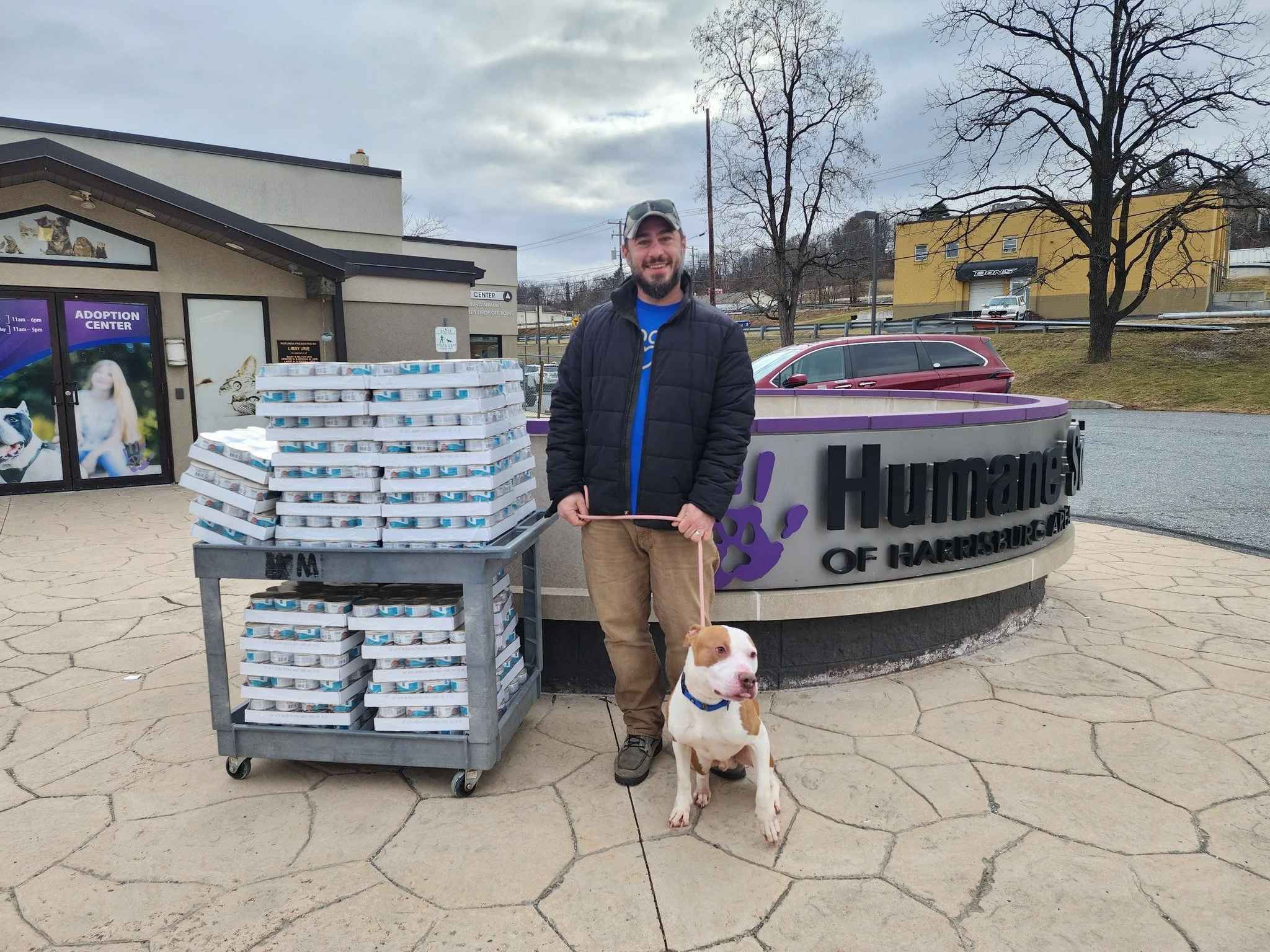 A man with a beard and cap, wearing a black jacket, stands outside Humane Society of Harrisburg with a brown and white dog on a leash. They are near a cart filled with cases of canned dog food. Behind them is a building with a sign for an adoption center, a yellow building, trees, and parked cars. The sky is overcast.