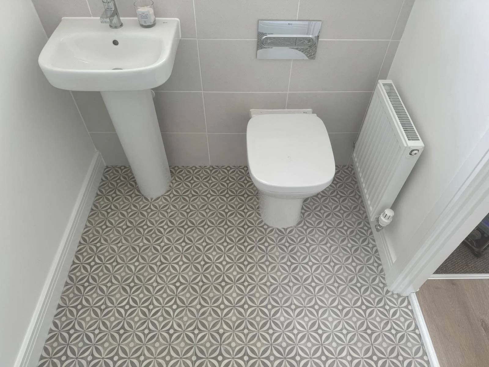 A small bathroom with a white pedestal sink, a white toilet, and a white radiator on the wall. The walls are tiled in gray and white, and the floor has a geometric gray and white pattern.