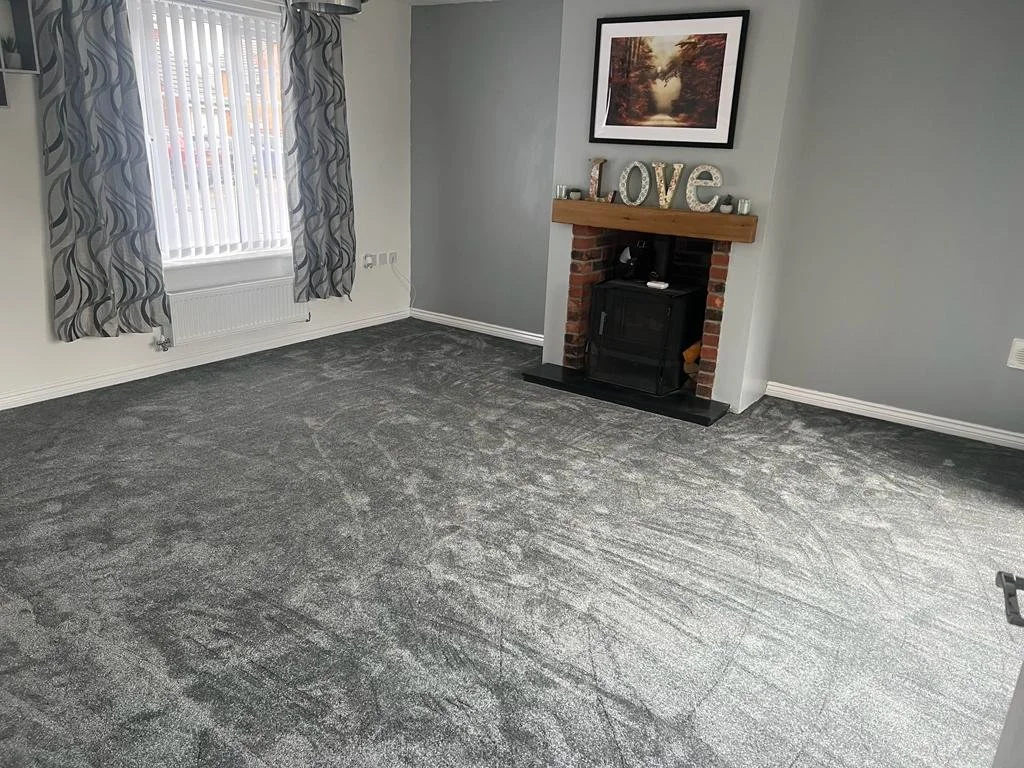 Empty living room with grey carpeting, a window with patterned curtains, and a fireplace with a wooden mantle decorated with a "LOVE" sign. A framed picture hangs above the fireplace.