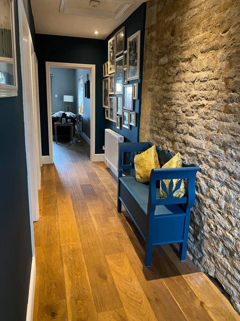 A narrow hallway with wooden flooring, blue walls, and a stone accent wall on the right. A small blue bench with two yellow pillows is against the stone wall, and a gallery of framed pictures decorates the blue wall. At the end of the hallway, an ope