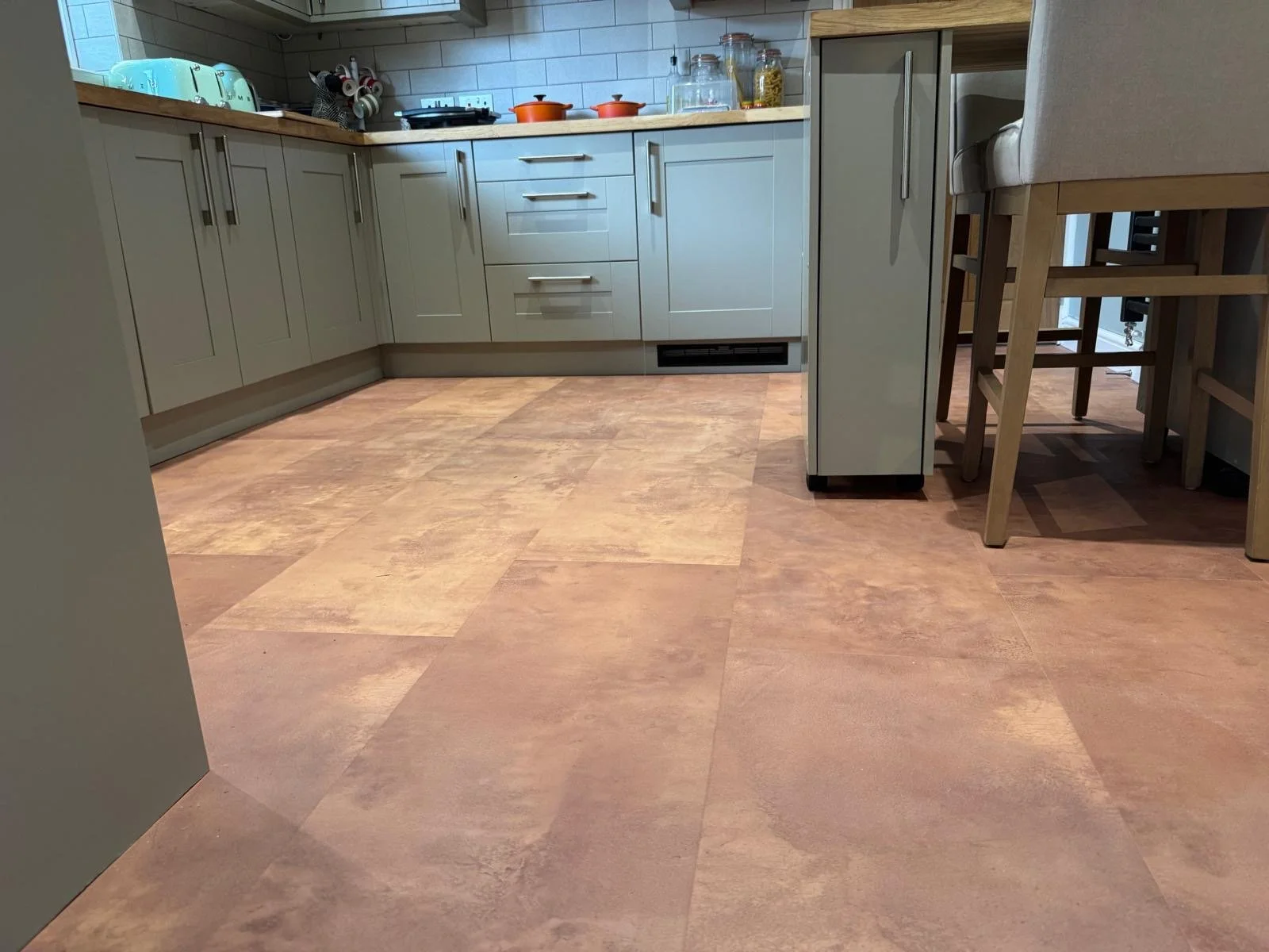 Modern kitchen with beige cabinets, wooden countertops, and pinkish-beige tile flooring. Items on the counter include a toaster, dishes, glass jars, and some orange pots on the stove.