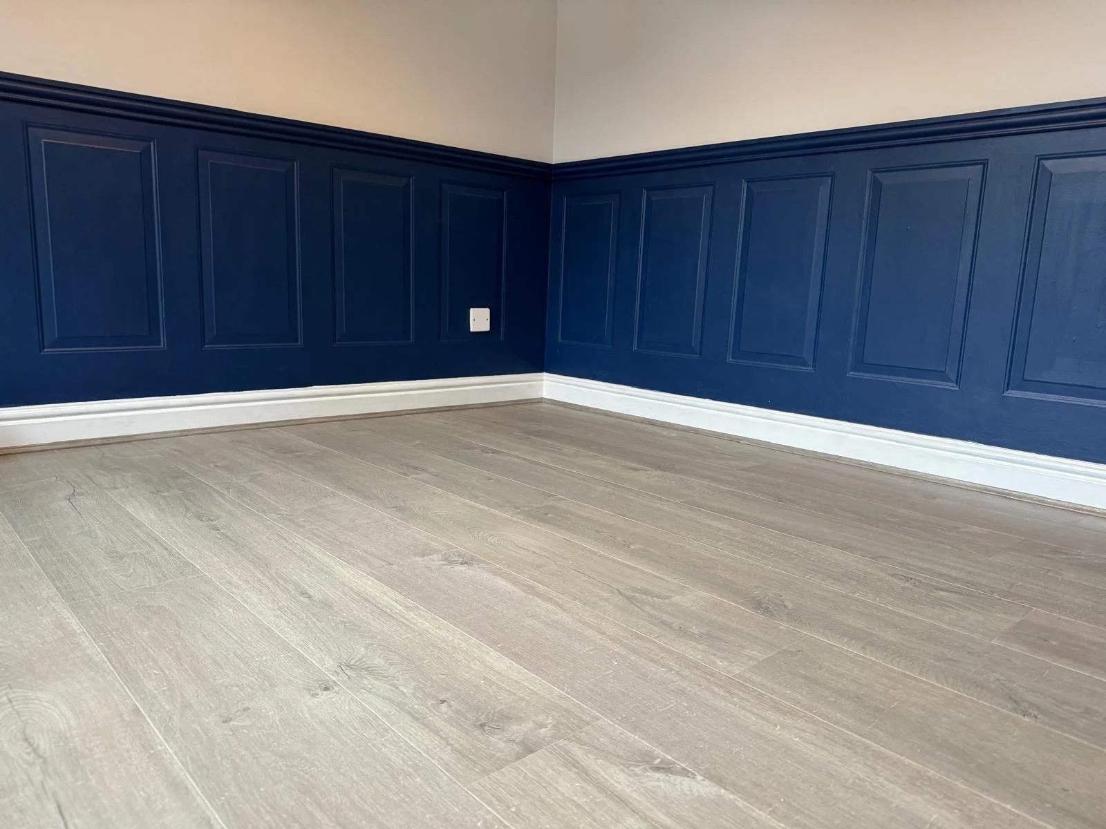 Empty room with beige wooden flooring, navy blue wall paneling, and a white baseboard.