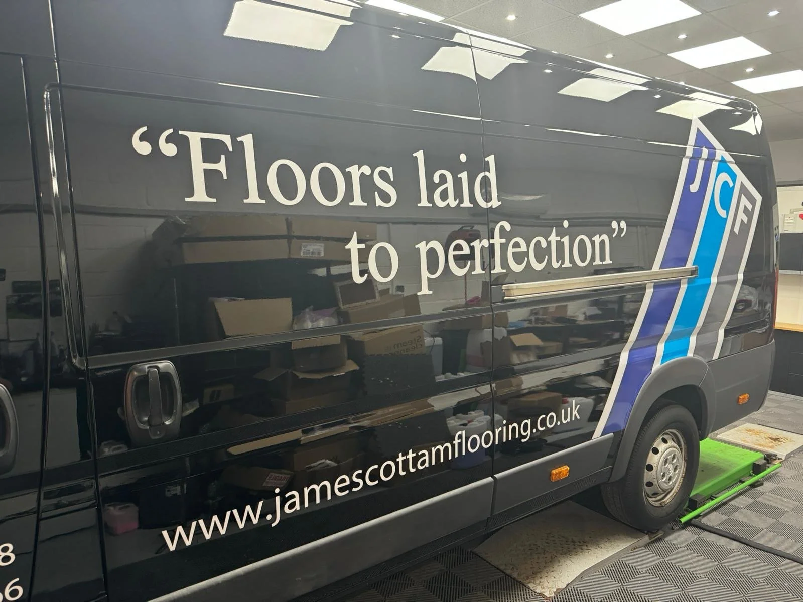 A black van with text advertising James Cottam Flooring, featuring the quote 'Floors laid to perfection' and a website address www.jamescottamflooring.co.uk, parked inside a room with a checkered floor and equipment underneath.