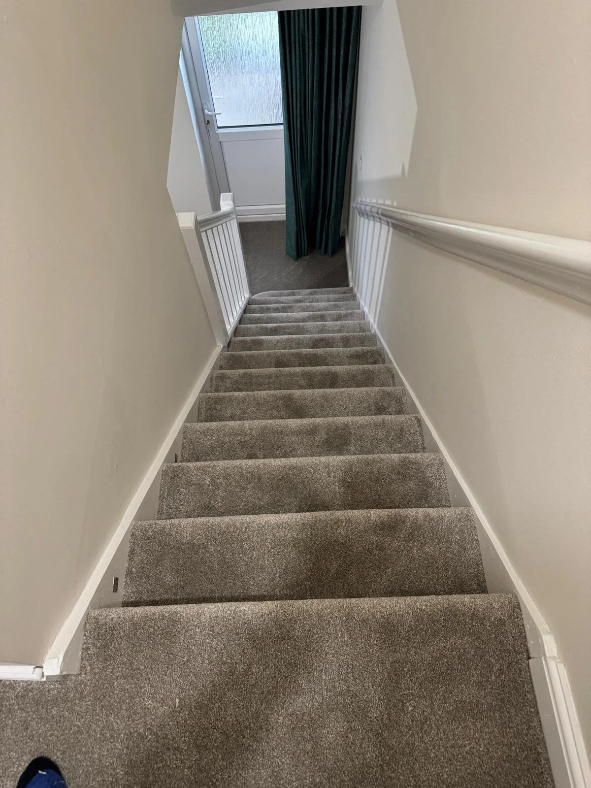 View looking down a carpeted staircase with white walls, white handrails, and a door with frosted glass at the bottom, with green curtains beside the door.