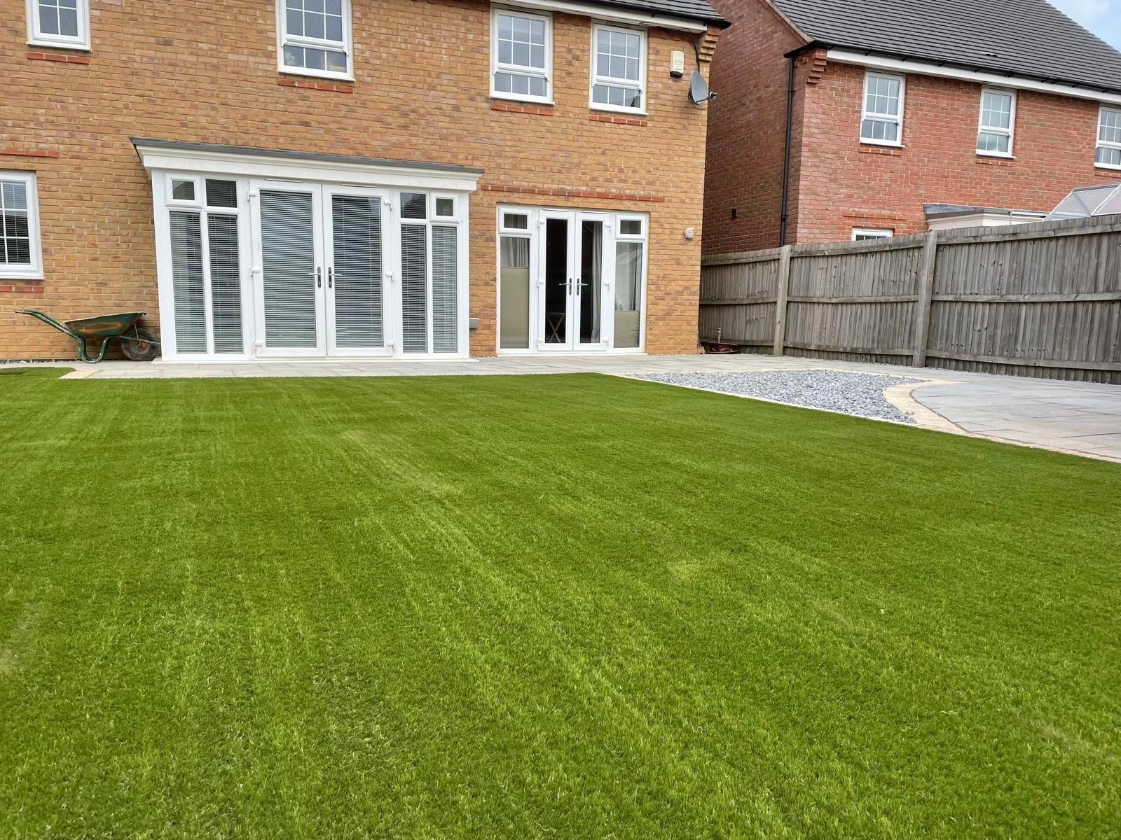 Backyard with well-manicured green lawn, patio area with stone tiles, wooden fence, and a brick house with large white-framed glass doors and windows.