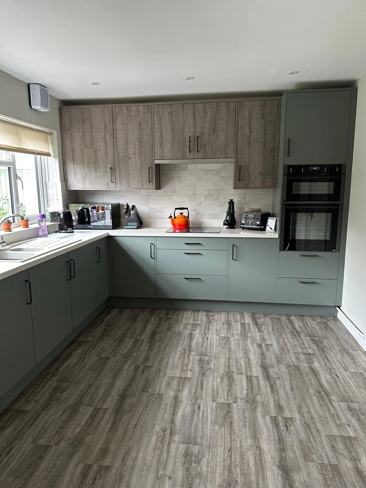 Contemporary kitchen with green cabinets, wooden upper cabinets, a window with a beige blind, and various small appliances on the white countertop, including a toaster, kettle, and coffee maker, with a wooden floor.