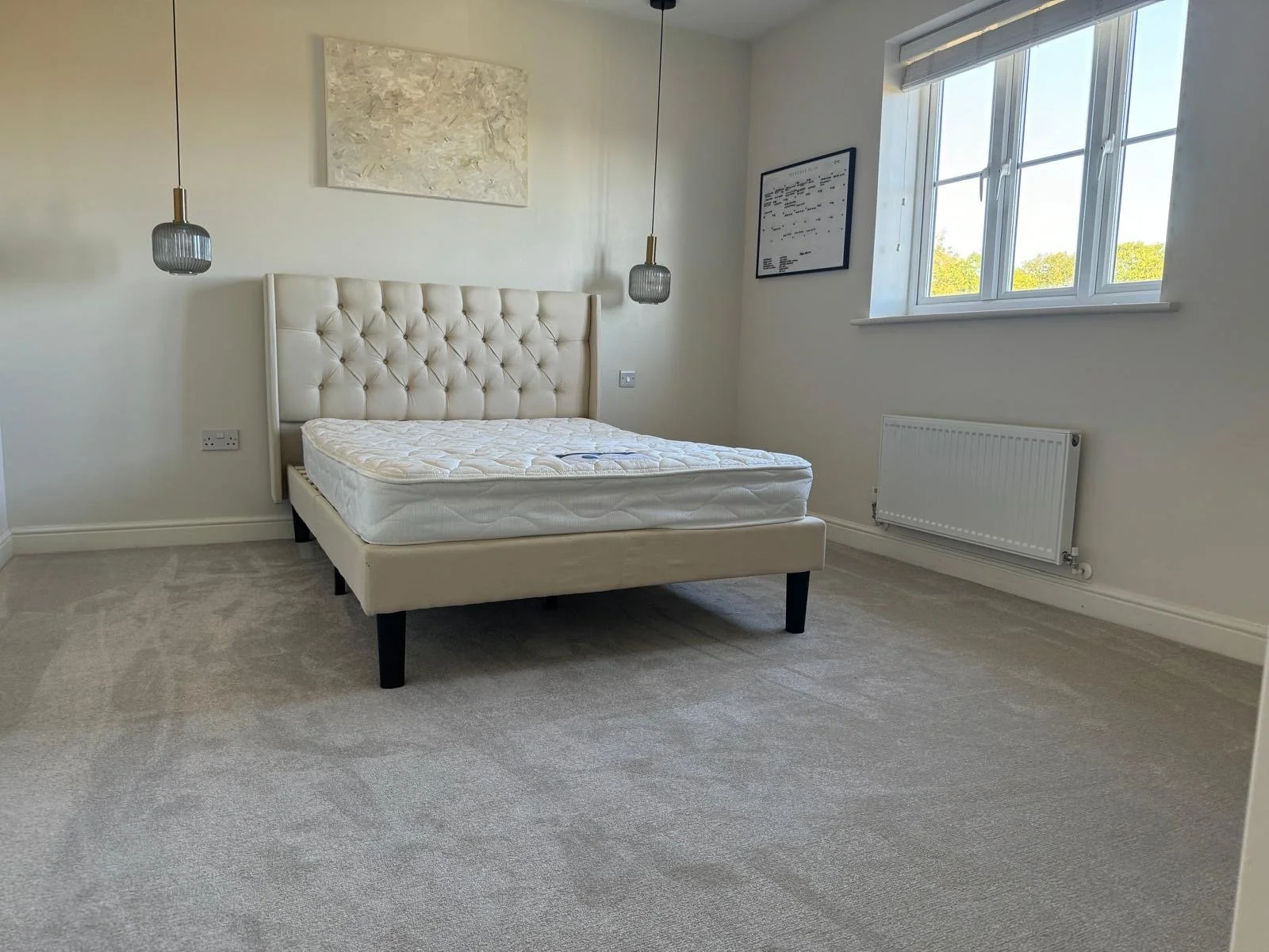 Empty bedroom with a large window, cream-colored carpet, a white tufted bed, two hanging pendant lights, and a white radiator