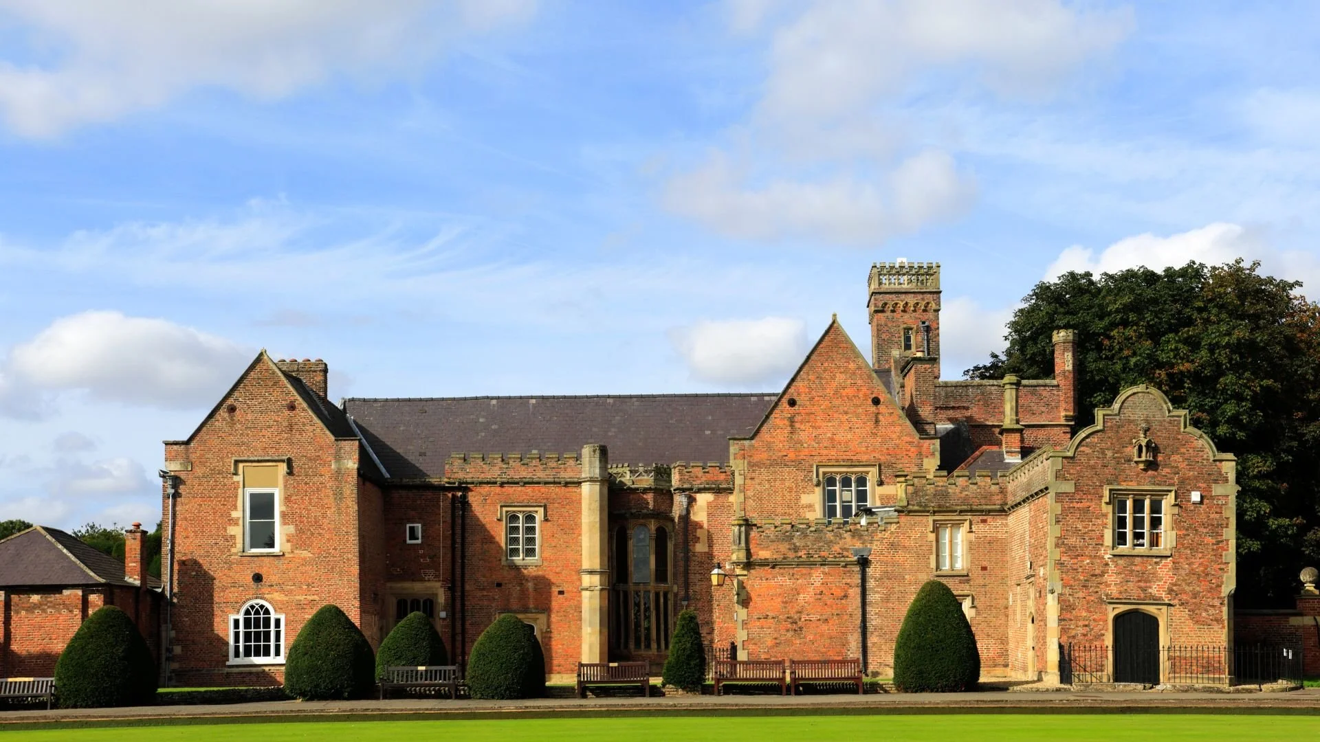 A historic brick building with multiple architectural features, including pointed gables, arched windows, a central tower, and decorative brickwork. The building is set against a partly cloudy sky with a well-maintained lawn and benches in the foreground.