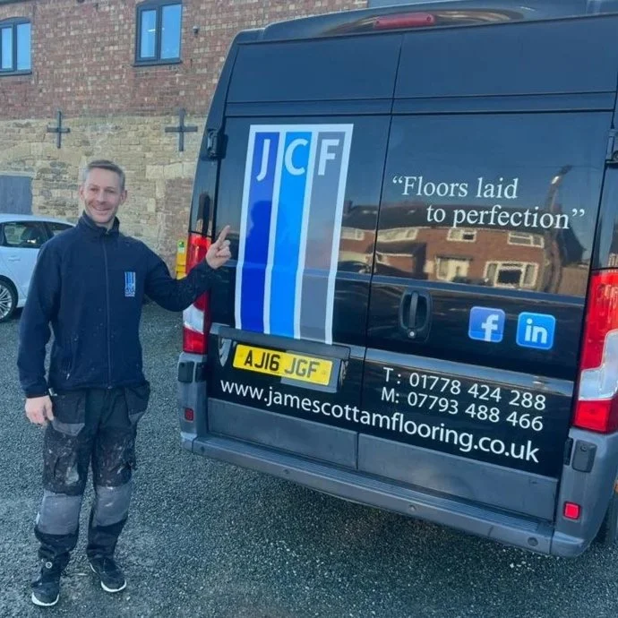 Man standing outdoors next to a black van with logo and contact information for James Scott Flooring, pointing at the logo.