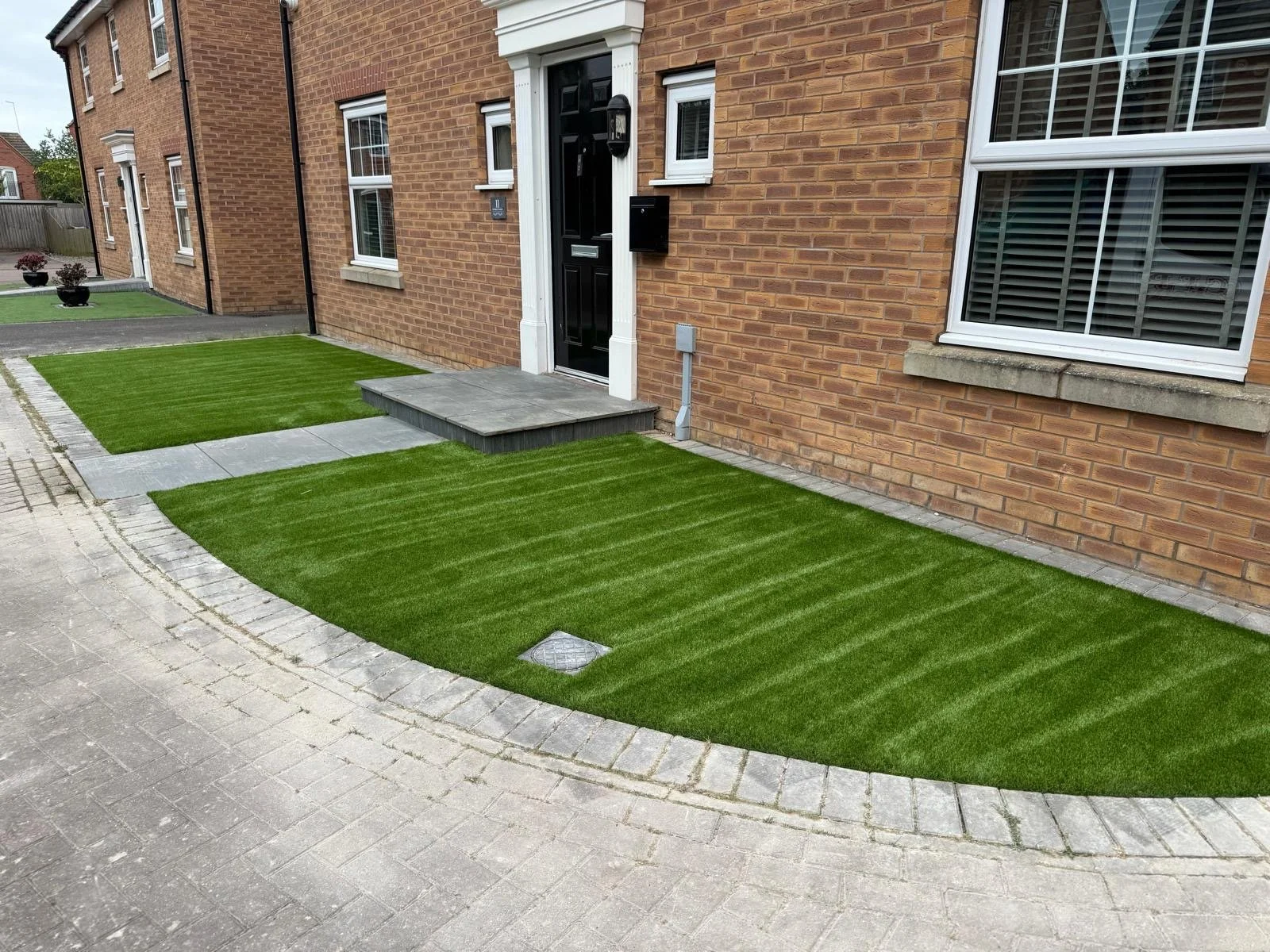 Well-maintained front yard with striped artificial grass, a paved stone pathway, and a brick house with a black front door and large window with white frame.